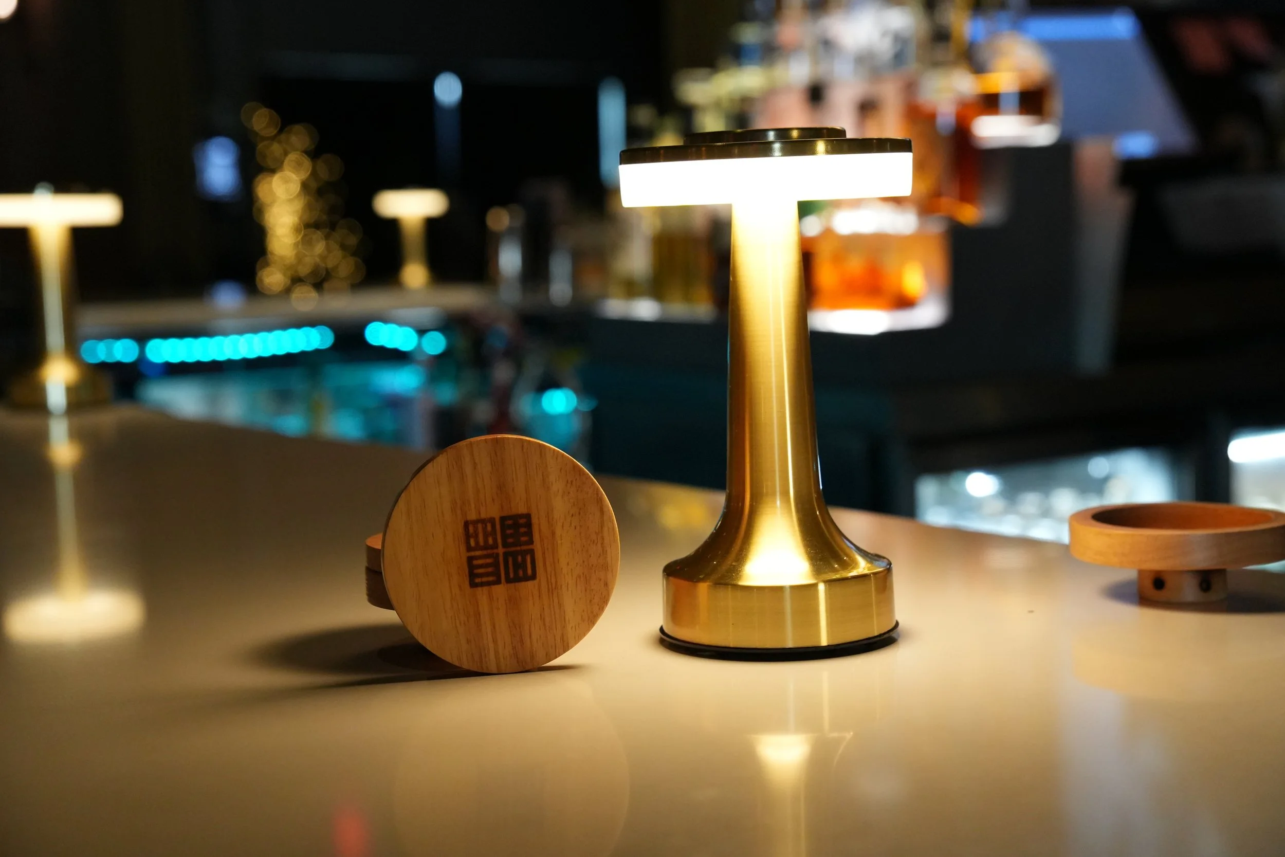 A gold-colored table lamp with a cylindrical white shade casting light on a table. In the background, blurred lights and decor are visible, creating a warm ambiance.