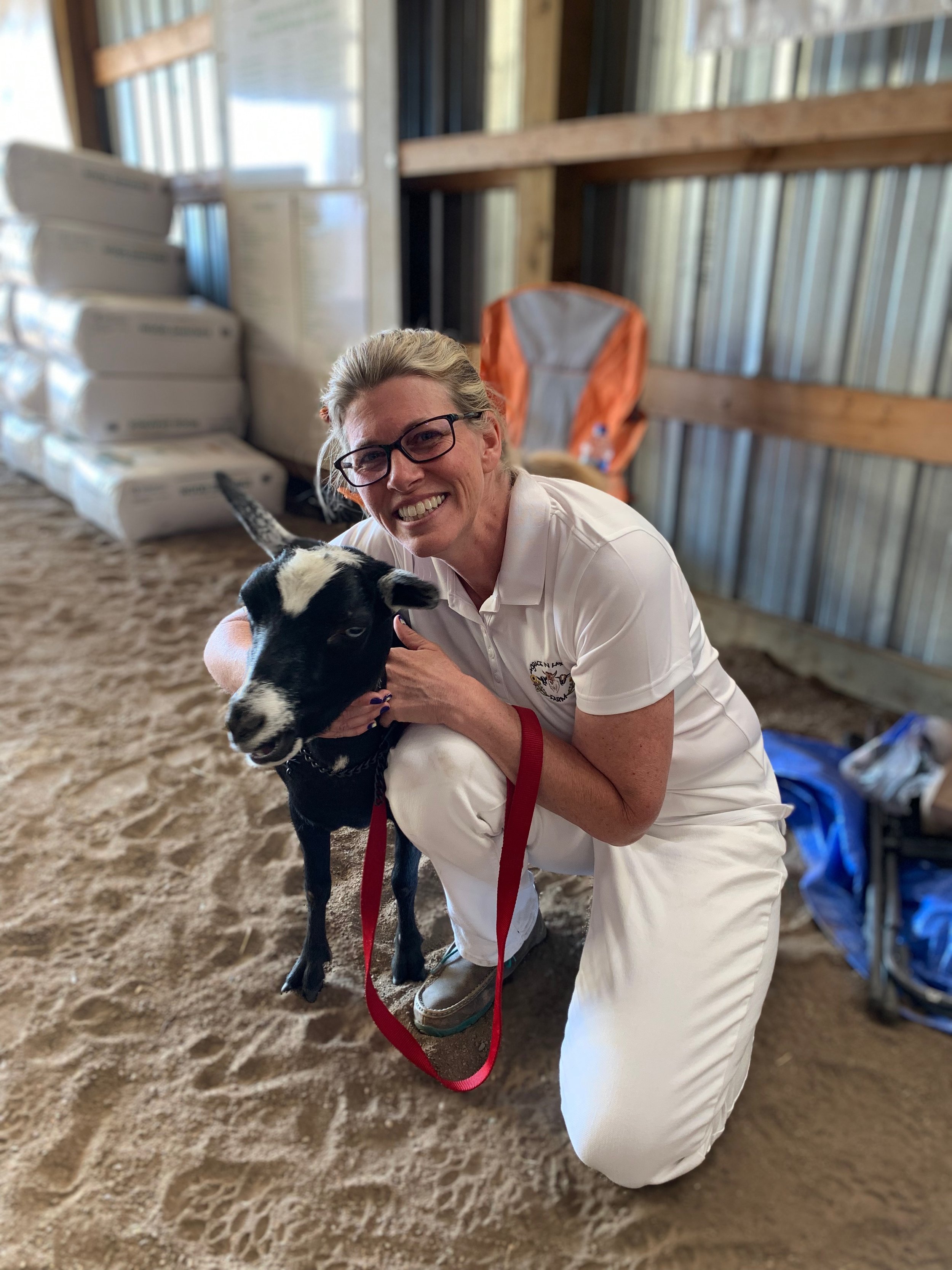 A woman with glasses kneeling on the ground in a barn, holding a black and white calf, smiling. Stacked boxes and metal walls are visible in the background.