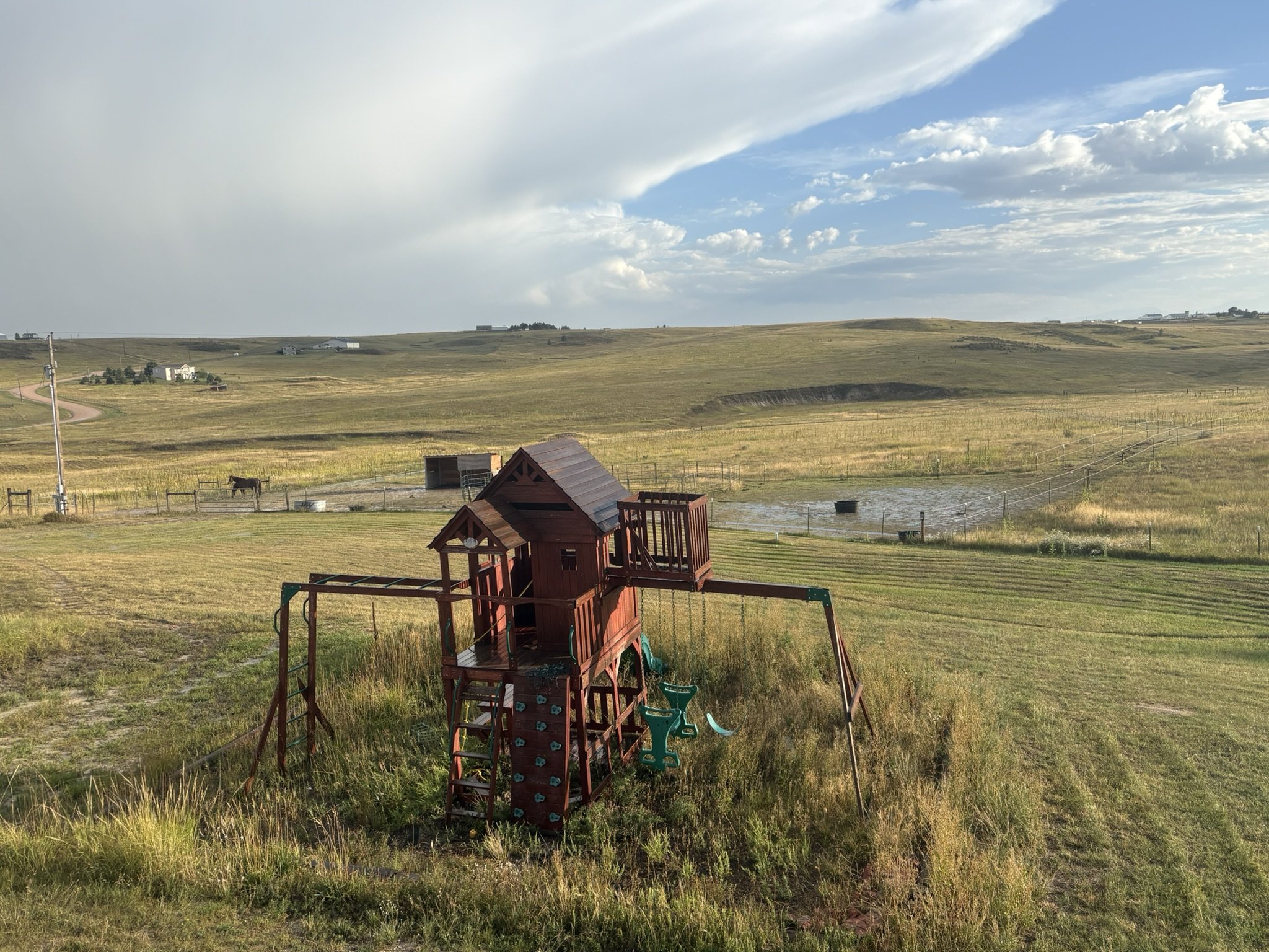 A rural landscape with green hills, a cloudy sky, and a red play structure in the foreground. There is a small pond and a horse grazing nearby, with a few houses visible in the distance.