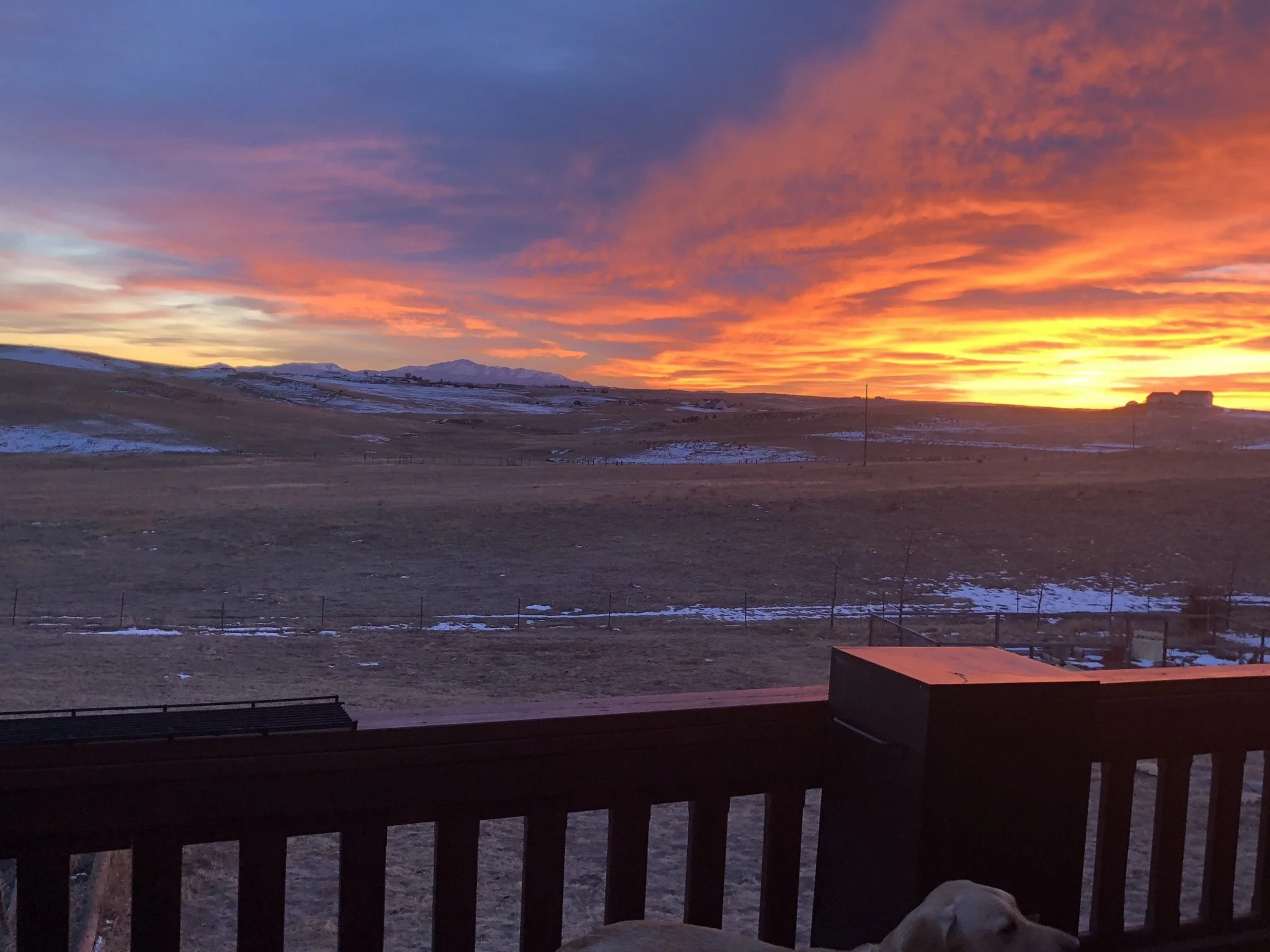 A sunset with colorful clouds over a rural landscape, seen from a porch or balcony with a wooden railing.