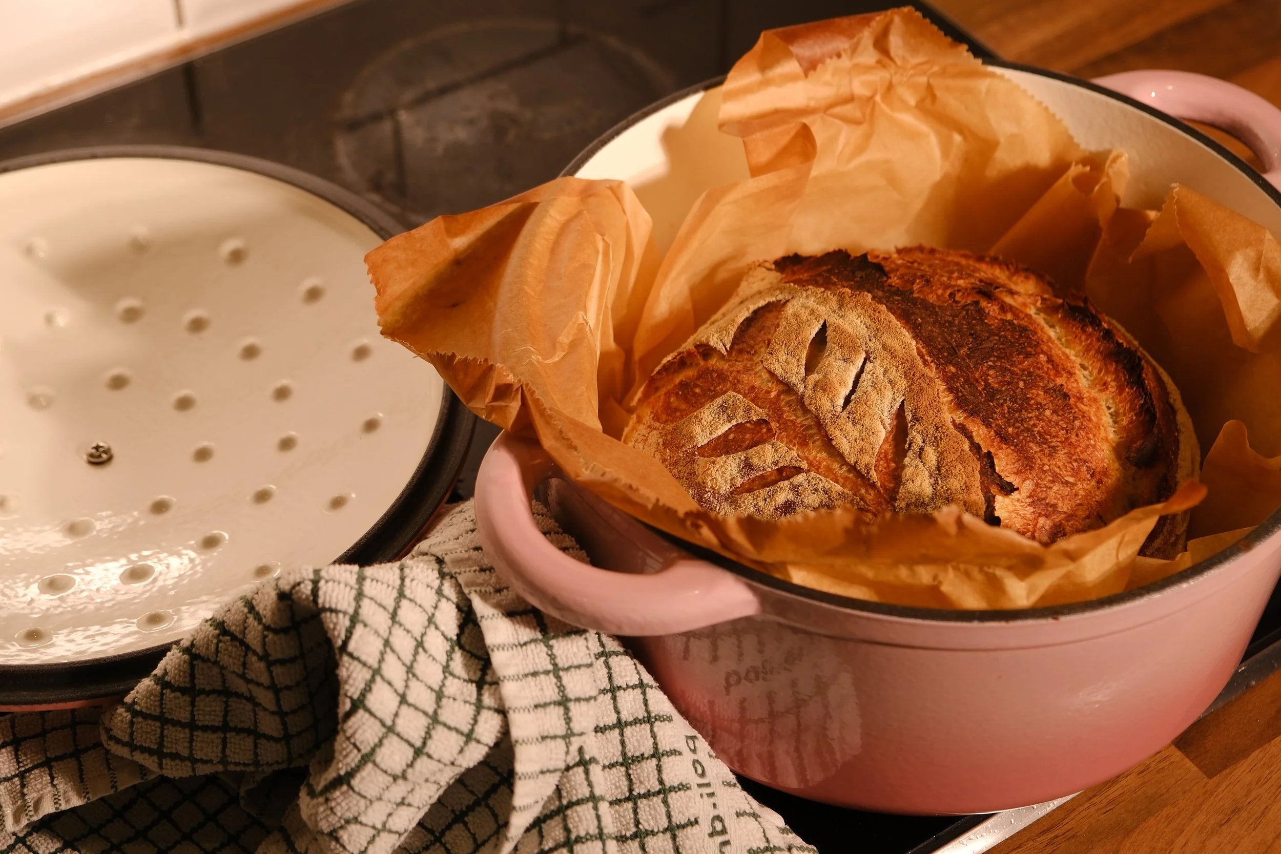 Freshly baked round loaf of rustic bread in a pink baking dish, lined with parchment paper, on a stovetop with a white dishcloth nearby.