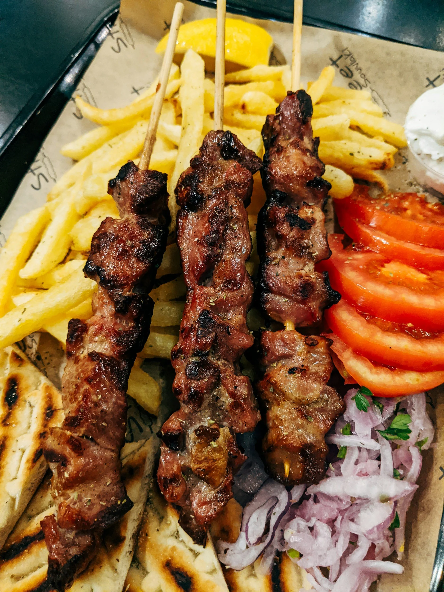 Three skewers of grilled meat with French fries, sliced tomatoes, chopped onions, and a small container of sour cream.