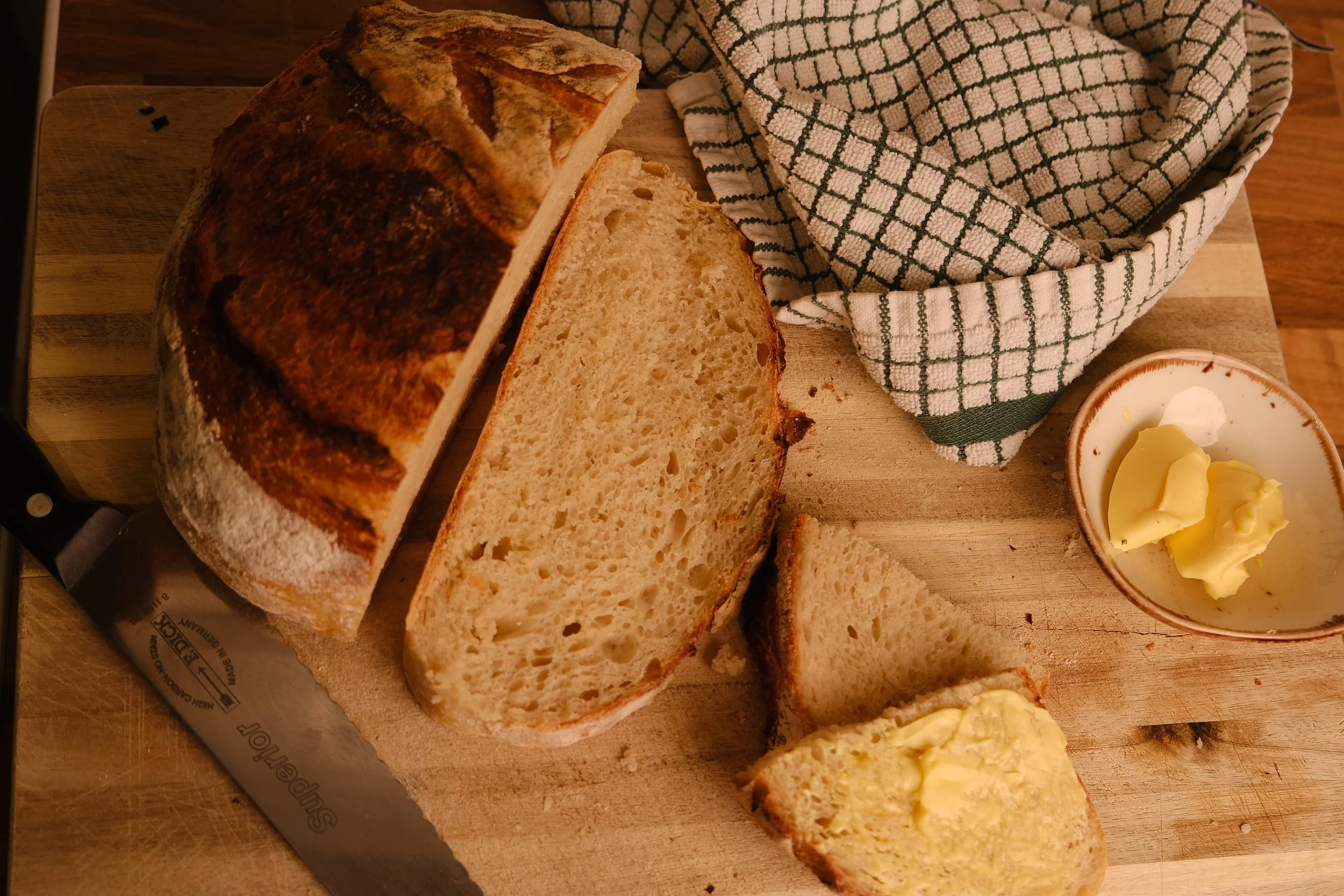Slices of freshly baked bread on a wooden cutting board, with butter in a small dish nearby, and a cloth napkin in the background.