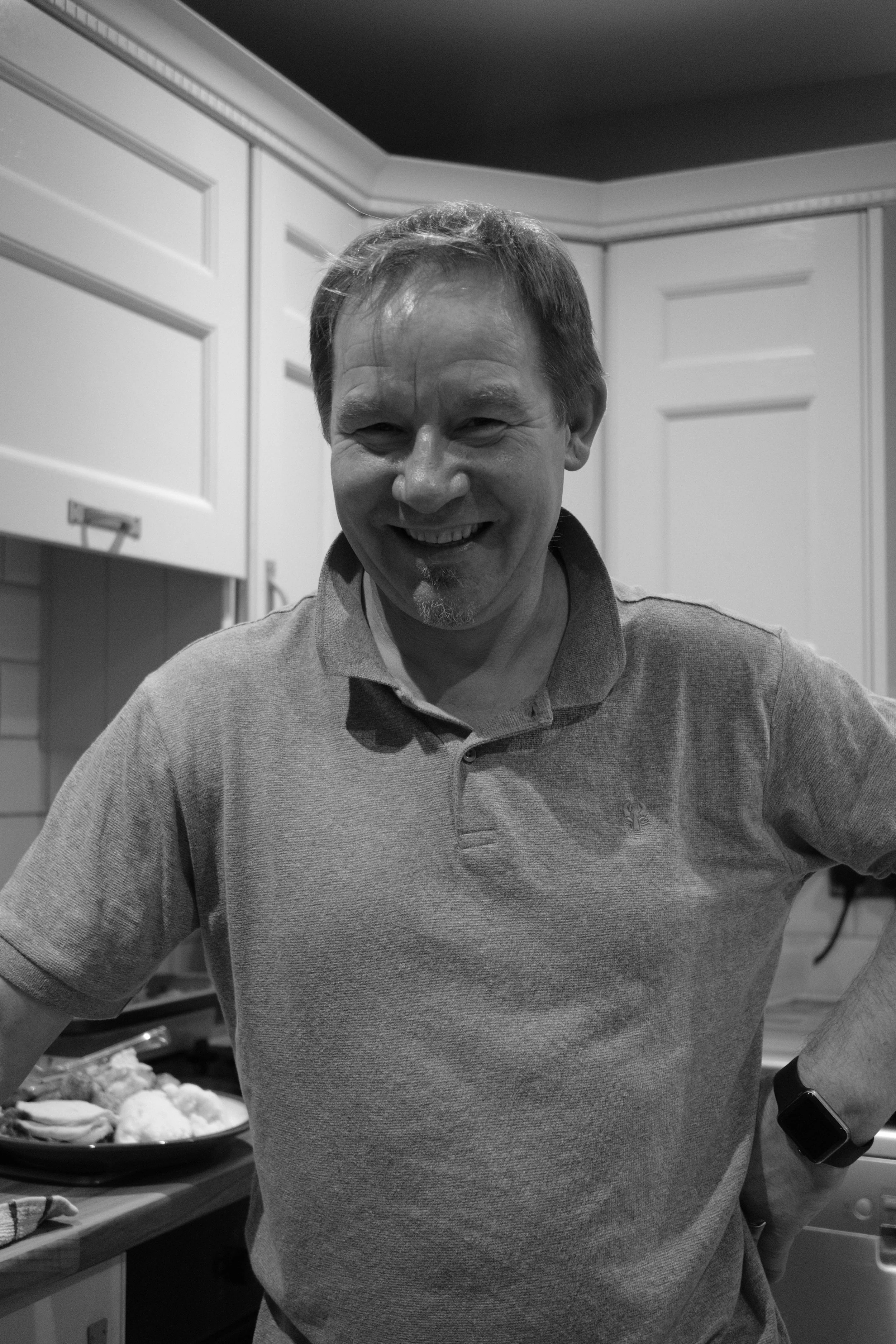 A chef off-duty smiling in a kitchen, wearing a polo shirt and a smartwatch.