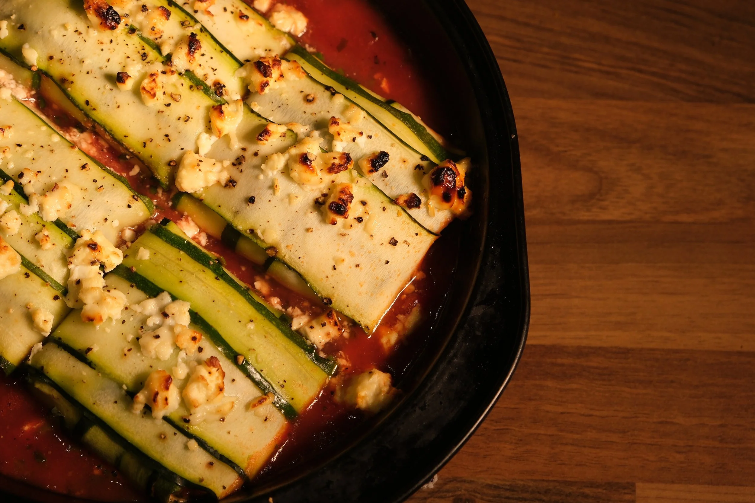 Zucchini noodles with crumbled cheese and pepper, baked in a tomato sauce in a black oven-safe dish on a wooden table.