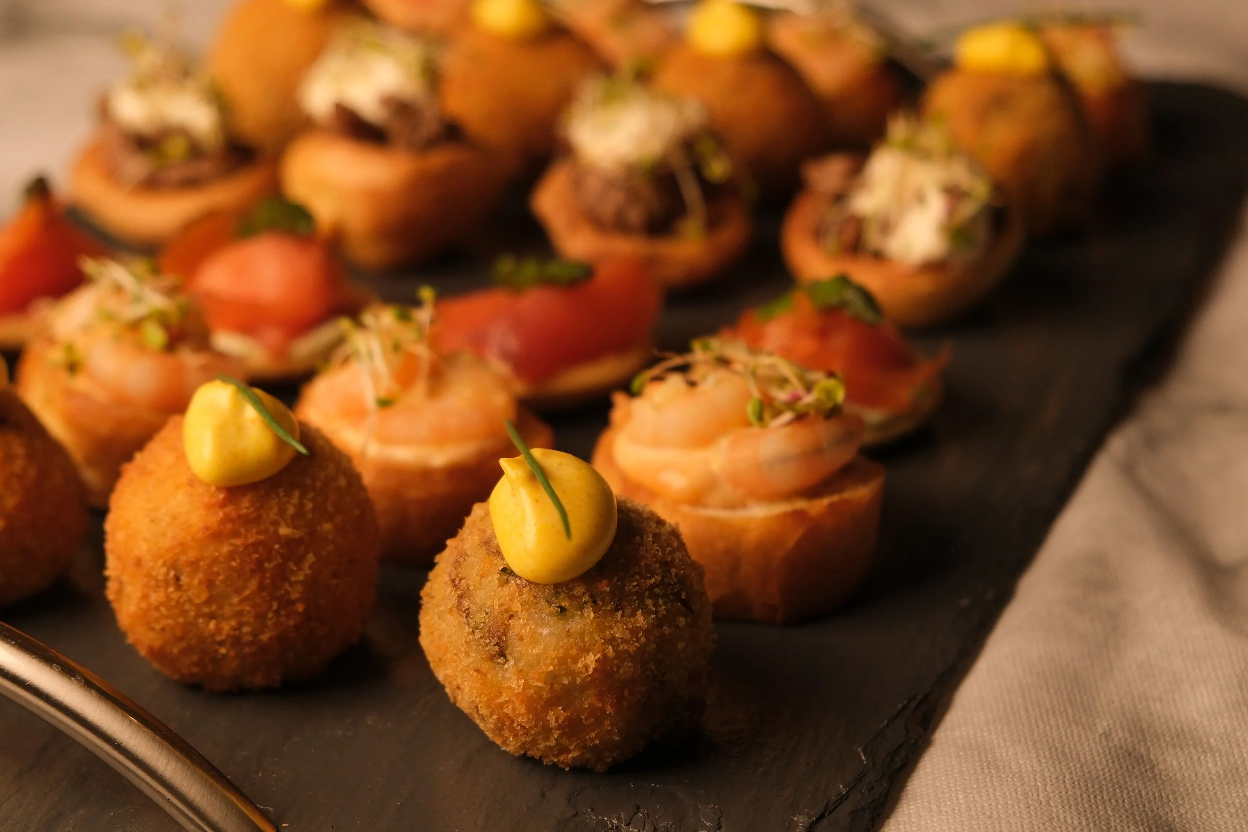 Assorted gourmet appetizers on a black serving tray, including croquettes topped with mustard and microgreens, and small seafood canapés with microgreens.