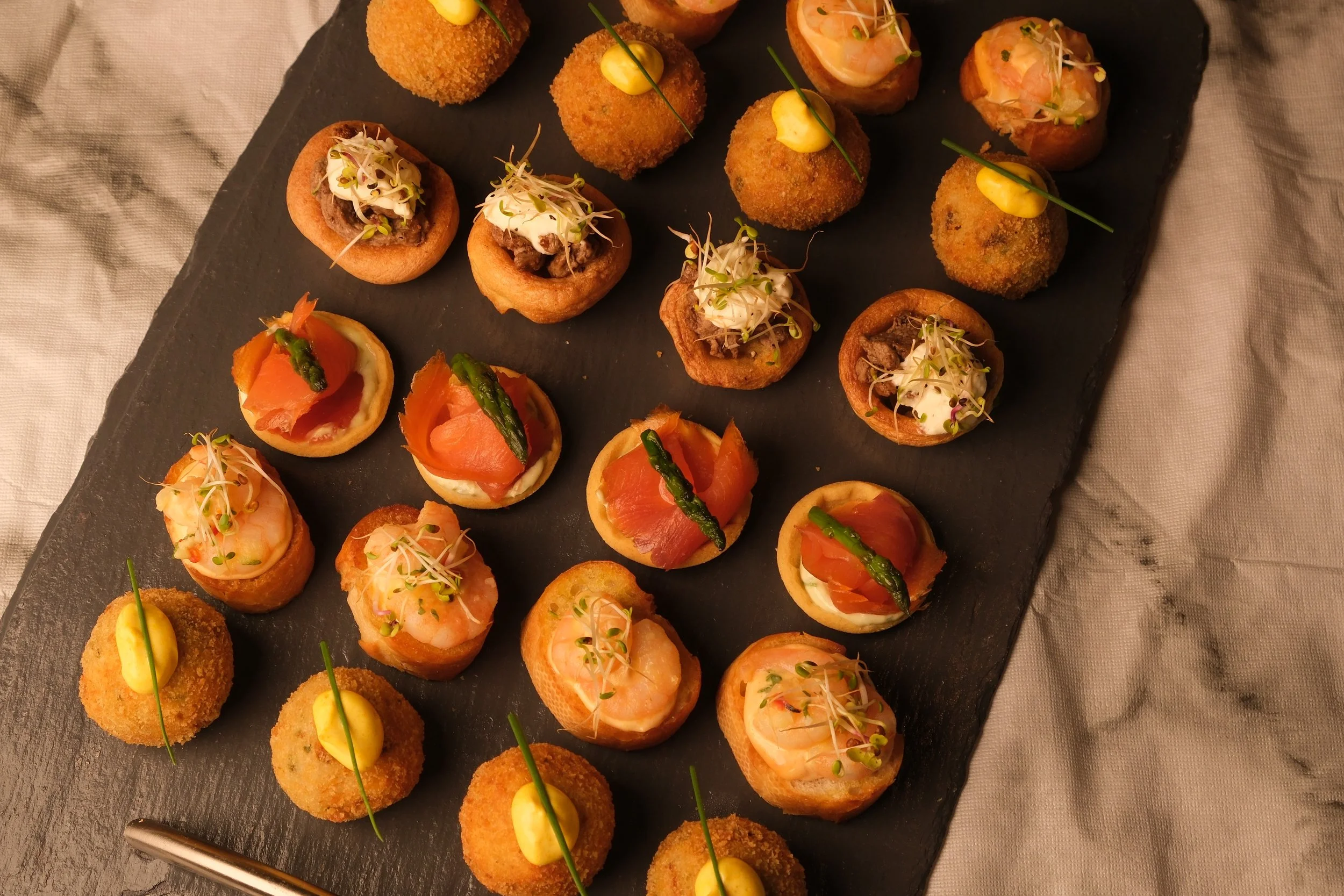 Assorted gourmet appetizers arranged on a black slate serving platter, including mini croquettes, smoked salmon canapés topped with asparagus, and other finger foods garnished with microgreens, presented on a textured gray tablecloth.