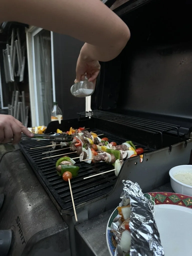 Person seasoning skewers of vegetables and meat on a charcoal grill at an outdoor barbecue.
