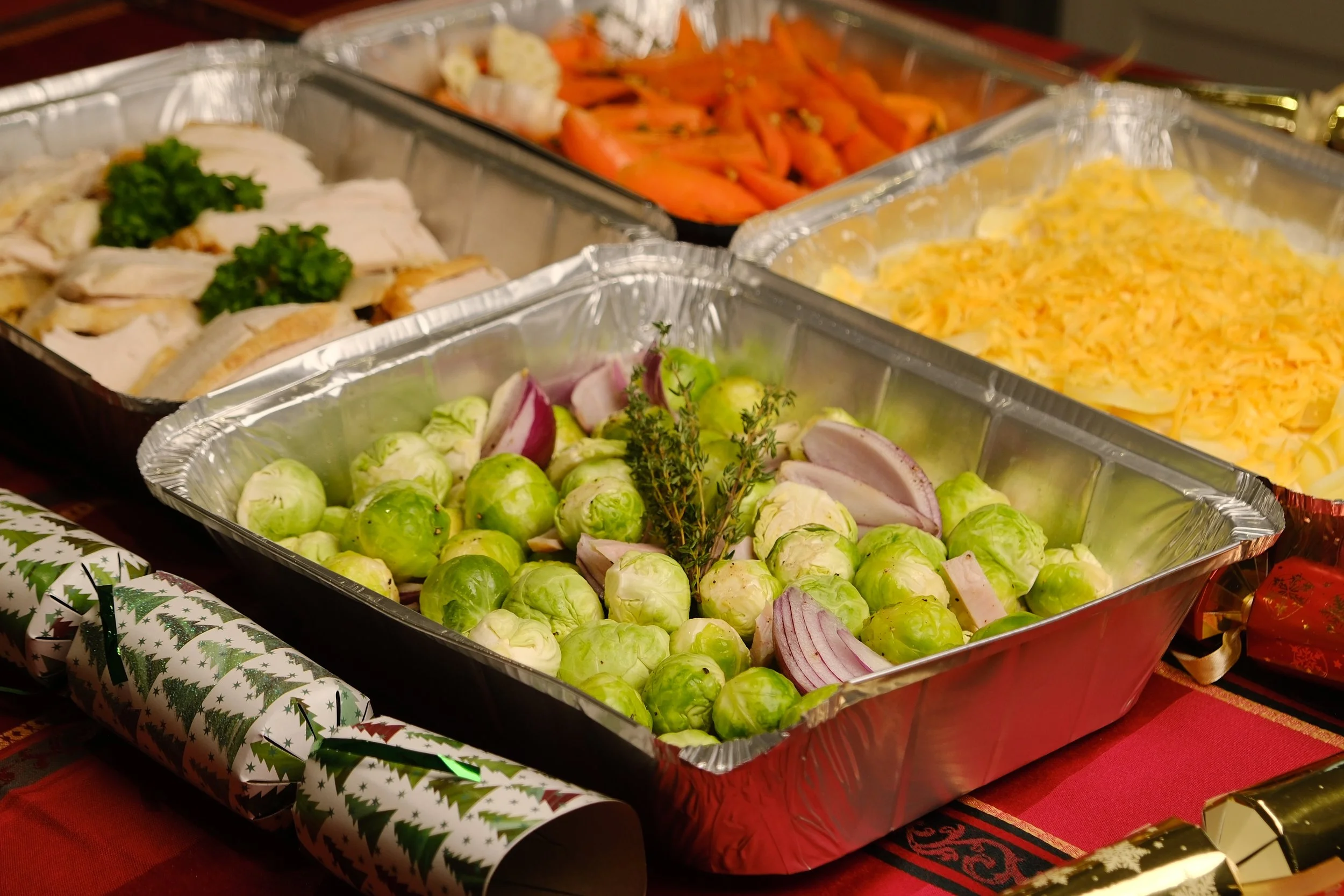 A festive holiday buffet with trays of Brussel sprouts, sliced red onions, shredded cheese, cooked carrots, and sliced turkey, decorated with Christmas wrapping paper and a red tablecloth.