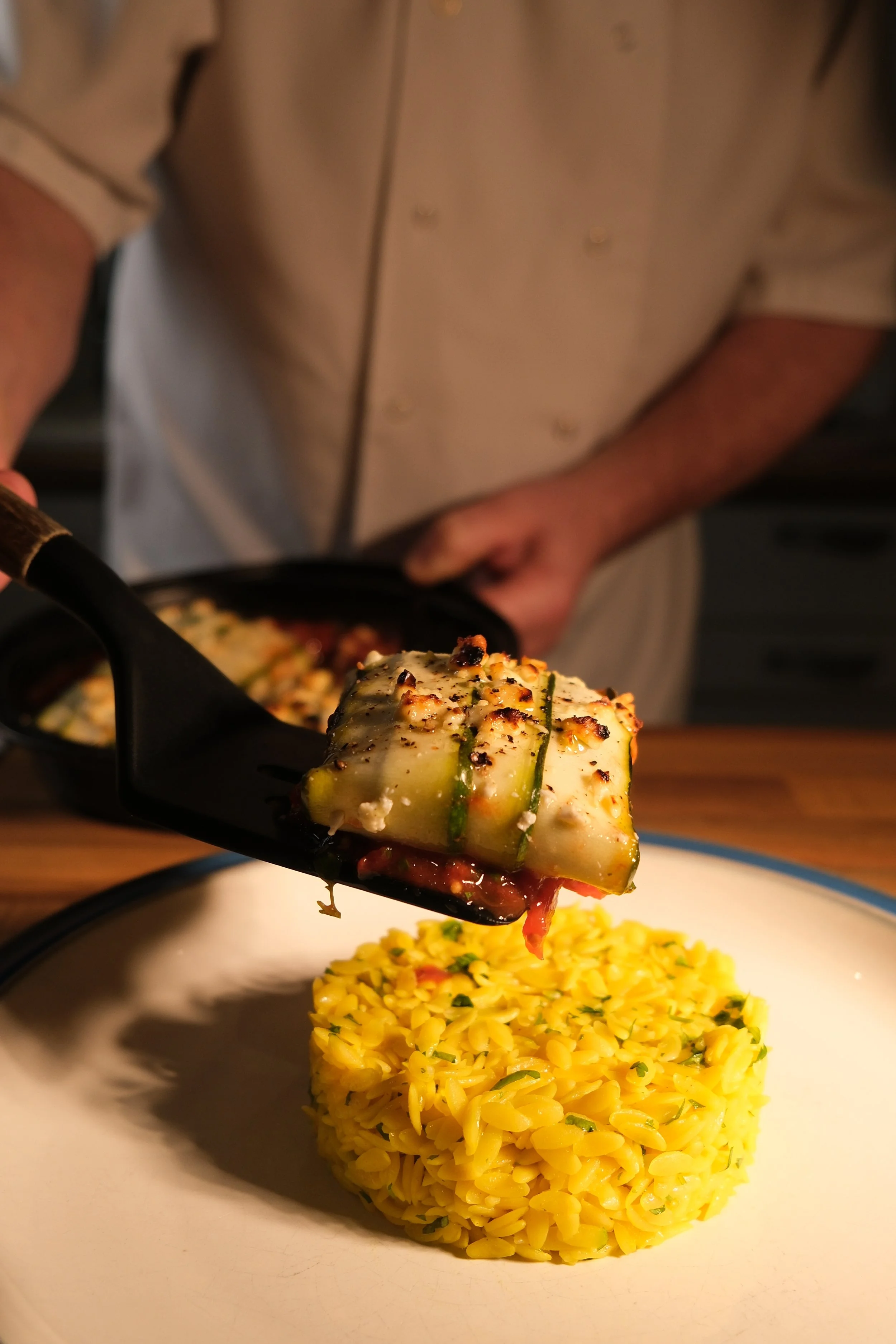 Close-up of a chef placing a grilled vegetable on a bed of yellow rice, with a focus on the food and chef in the background.