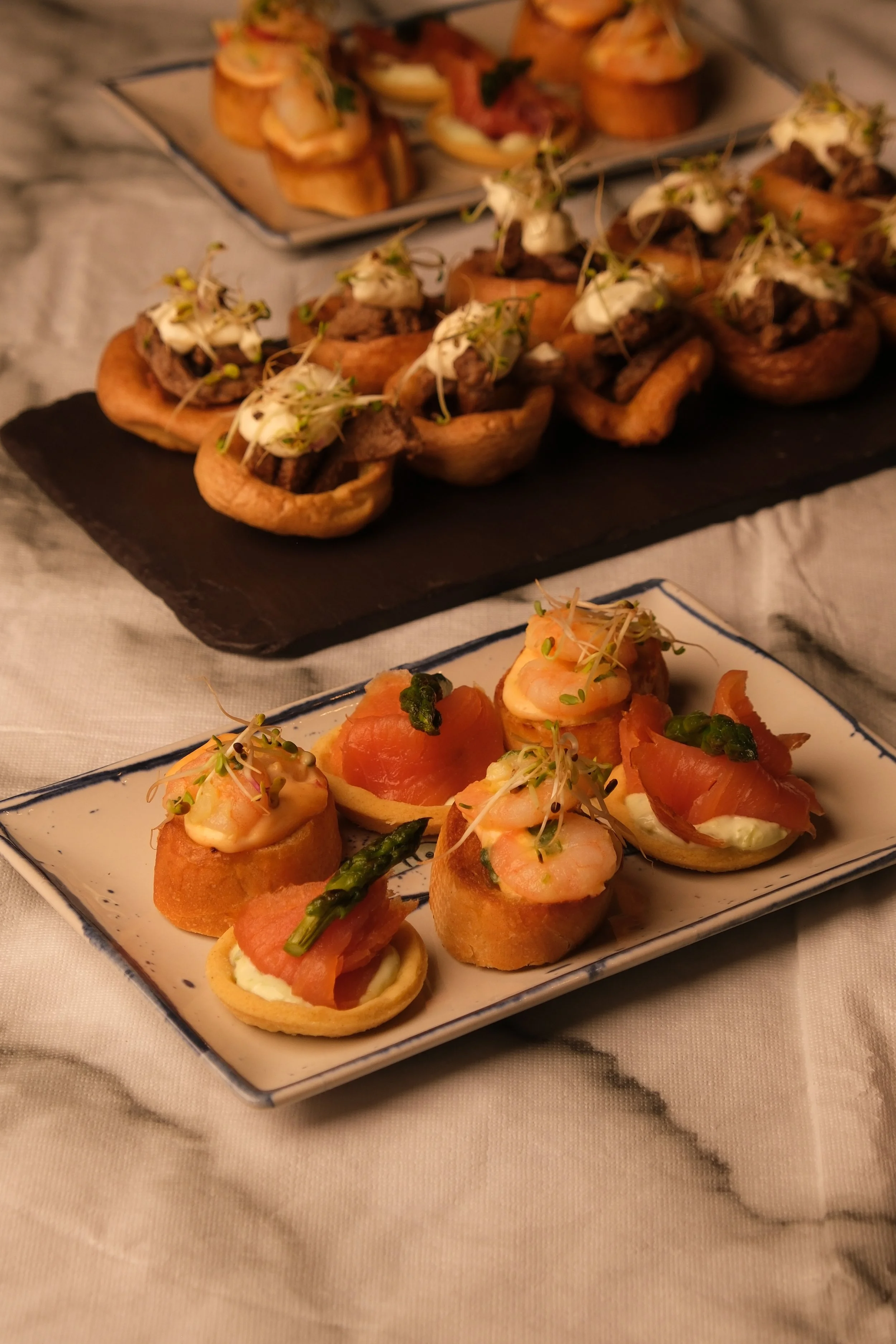 Appetizer platter featuring shrimp and smoked salmon on small toasts, garnished with microgreens and asparagus, with a variety of savory stuffed pastries in the background on a slate and a patterned ceramic plate.
