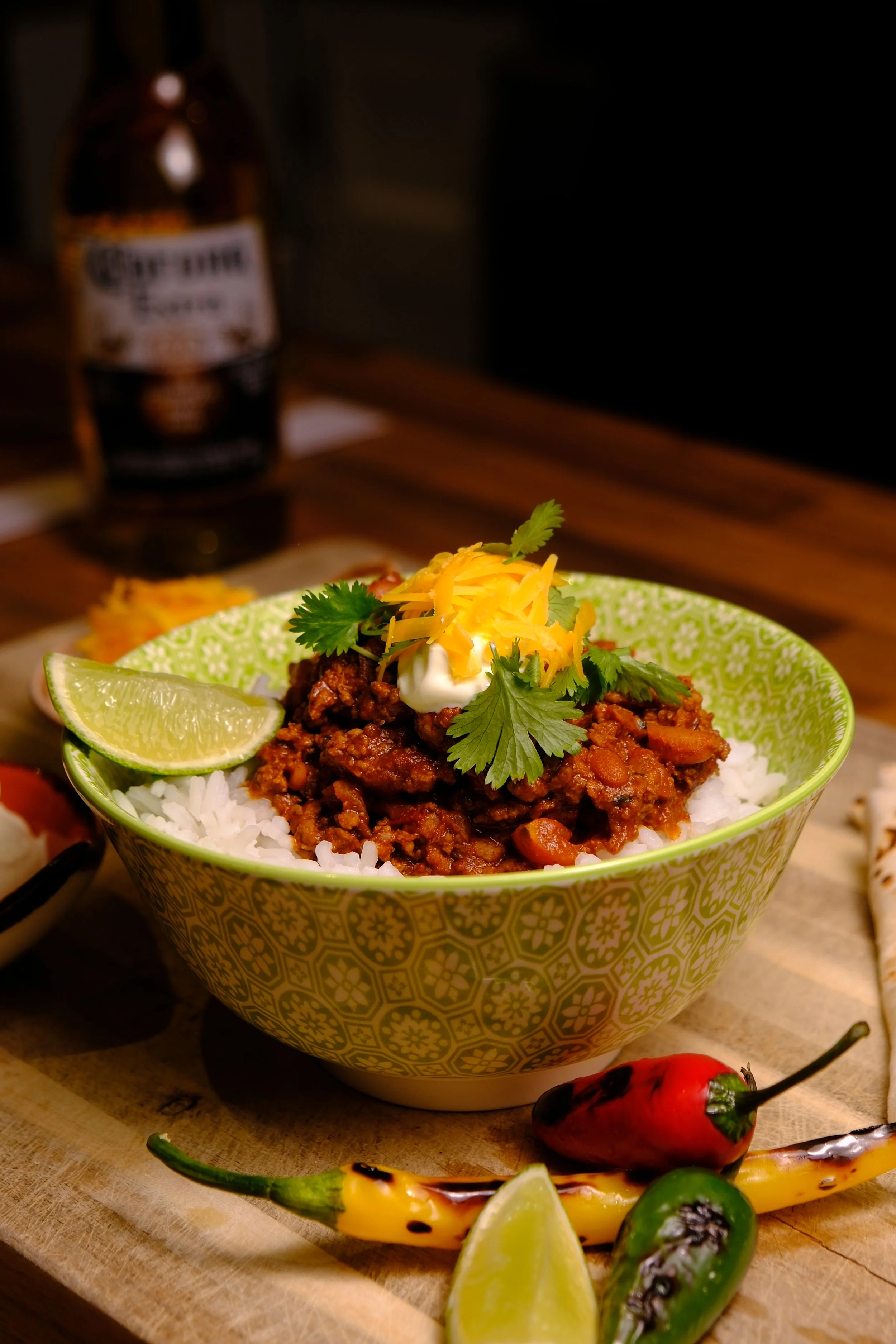 Bowl of chili served over white rice, garnished with shredded cheese, cilantro, and a lime wedge, with colorful chili peppers around it.