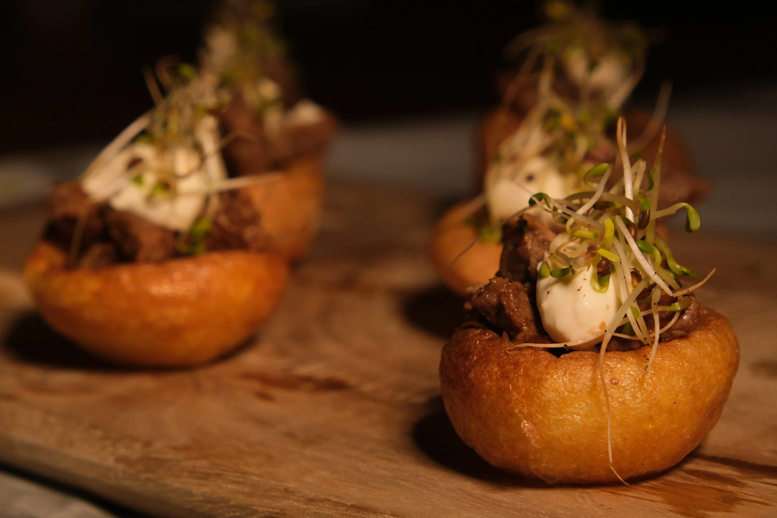 Miniature stuffed potatoes topped with sour cream, sprouts, and small pieces of cooked meat on a wooden surface.