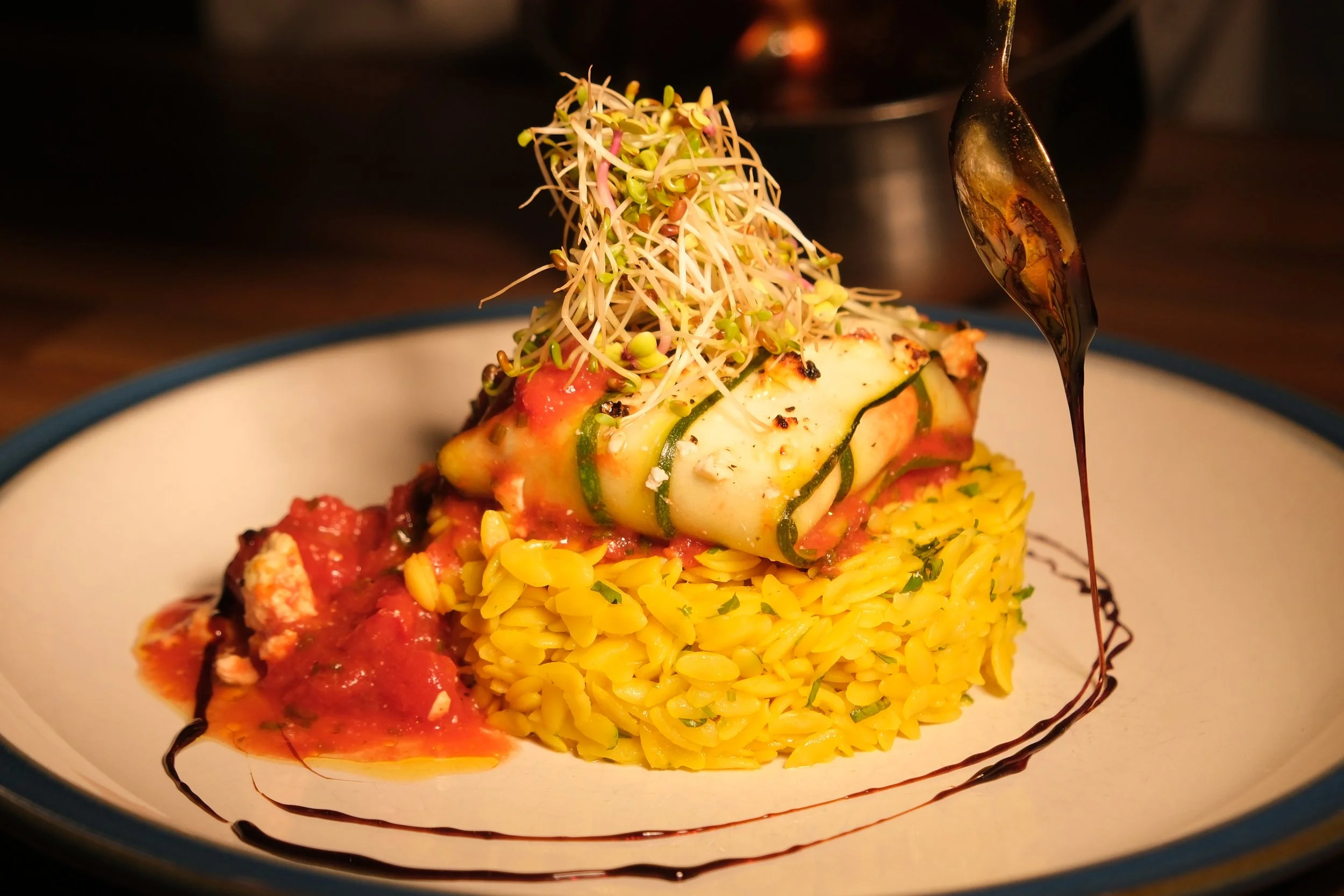 A plated dish of yellow rice topped with zucchini wrapped in cheese, garnished with microgreens, with tomato sauce and balsamic glaze drizzled around.