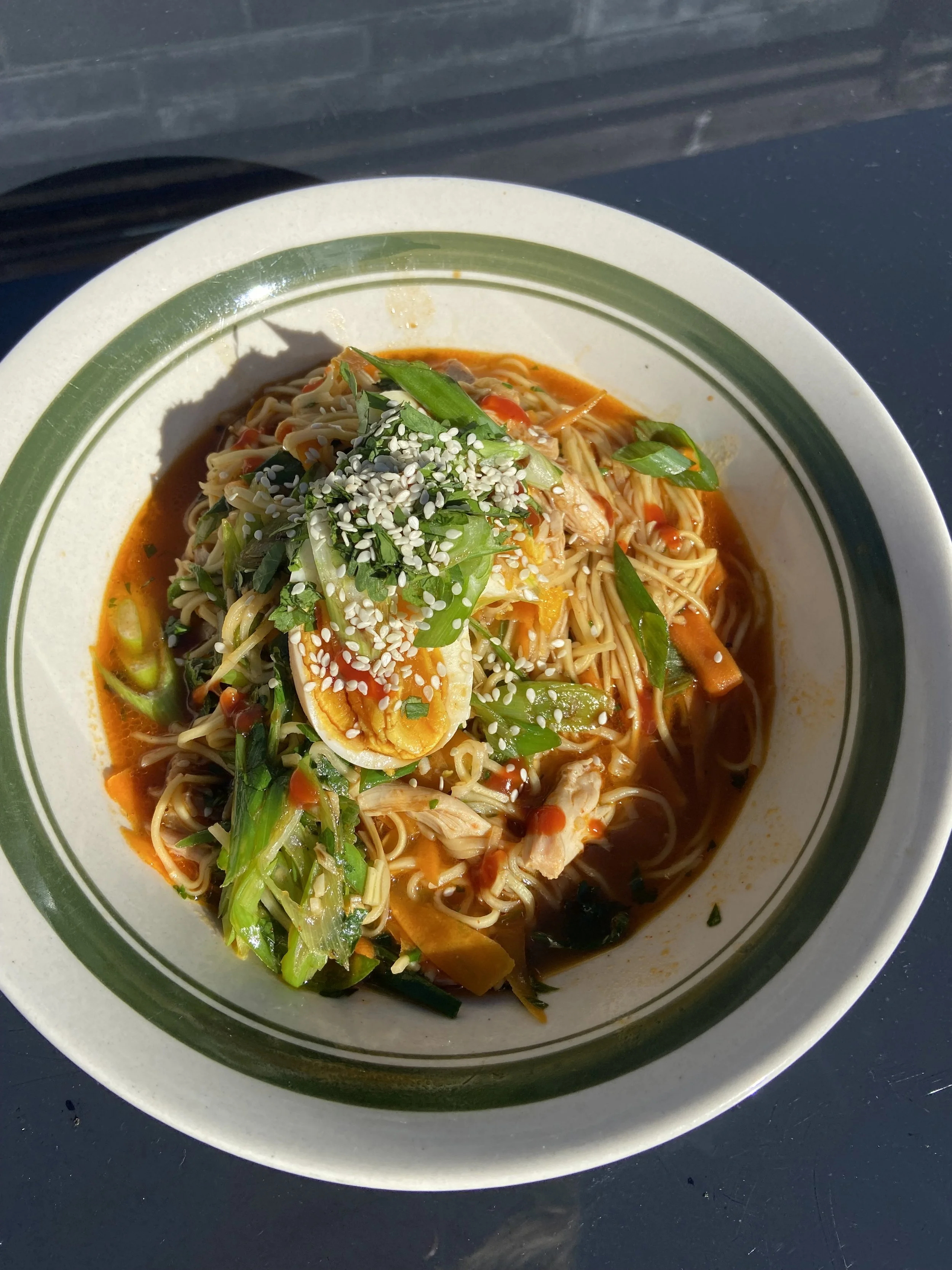 A bowl of Asian noodle soup with vegetables, a boiled egg, and sesame seeds, on a dark surface.