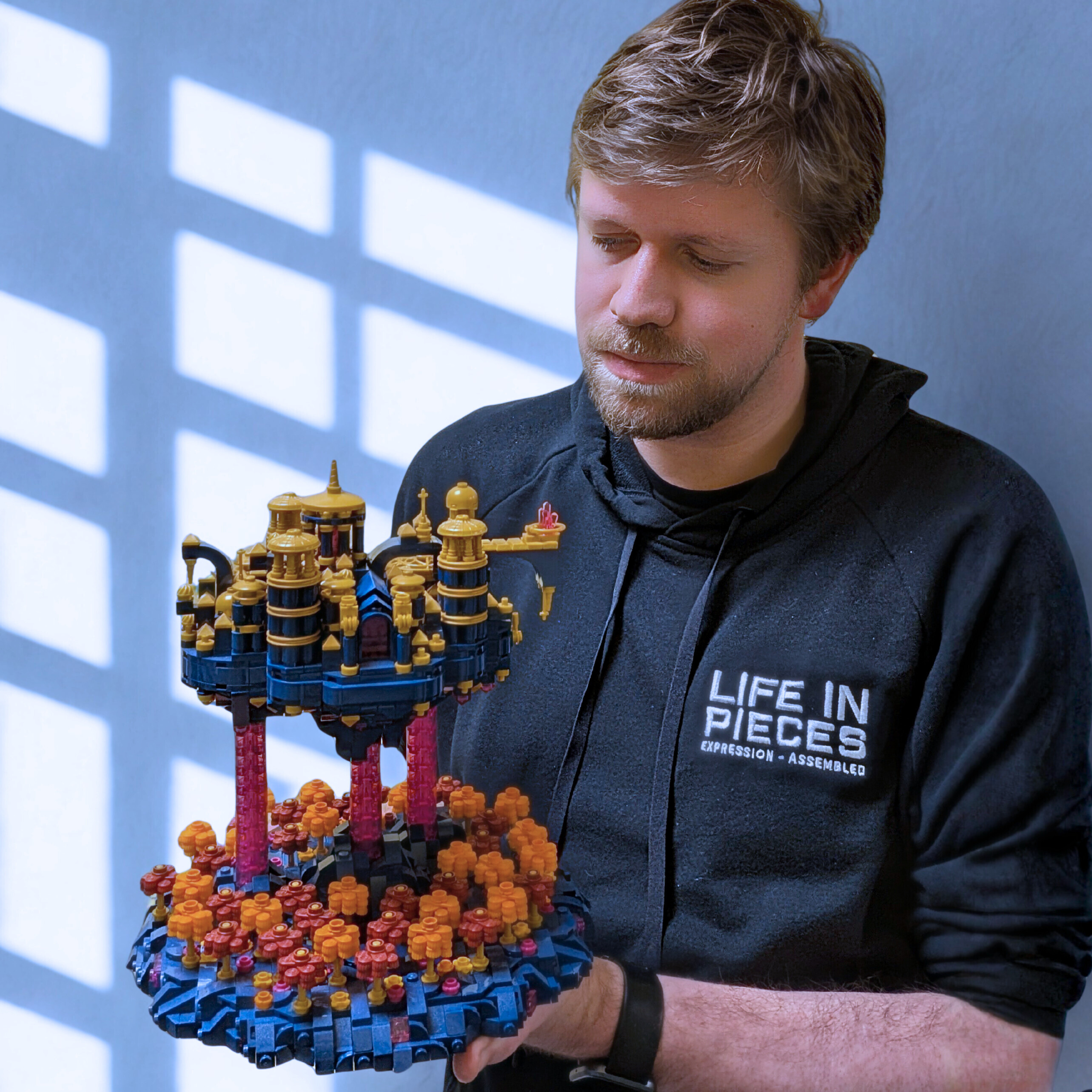 A man with light brown hair and a beard wearing a black hoodie with the words "LIFE IN PIECES EXPRESSION - ASSEMBLED" holds a large LEGO model. The model has multiple towers and is built on a round base with colorful LEGO pieces, resembling a futuristic floating city