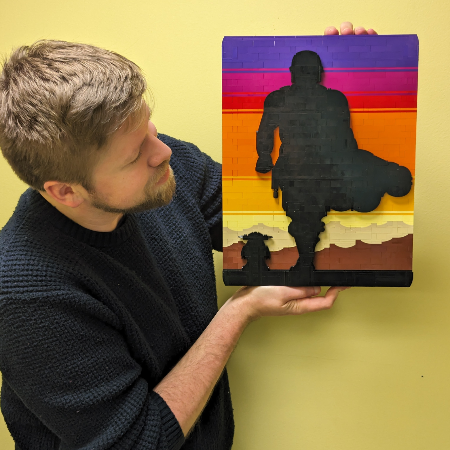 A man with light skin and a beard holding a colorful wooden sunset art piece featuring a silhouette of a soldier. The man is looking at the art, which is set against a yellow wall.