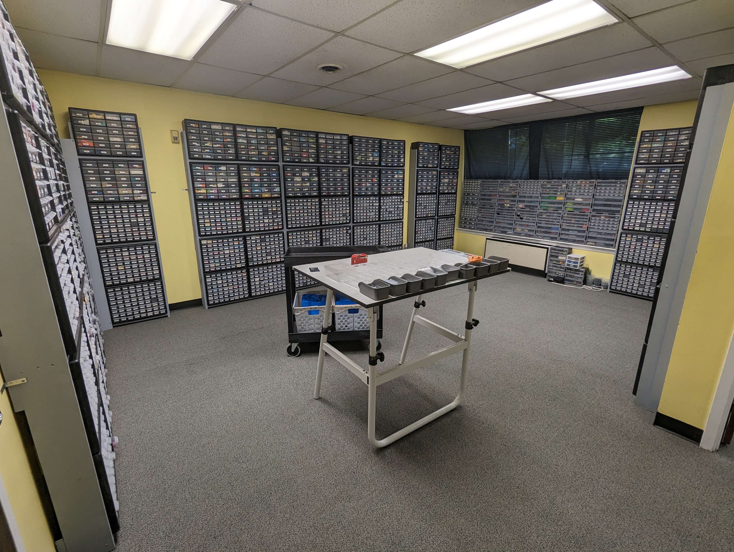 Room filled with black storage cabinets containing small labeled drawers, a table with sorting compartments, and a window with black blinds.