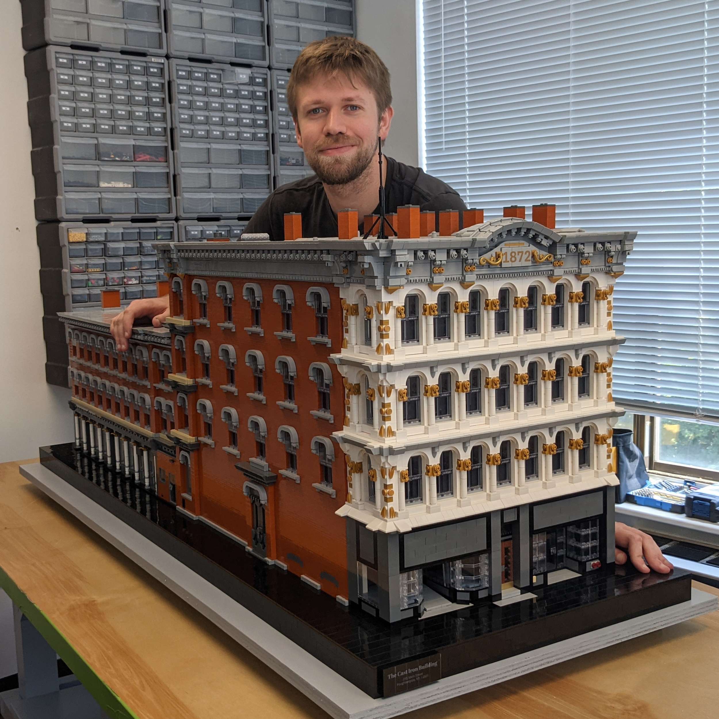 A man with a beard and short hair poses behind a large detailed LEGO building on a wooden table. The LEGO structure features multiple floors, decorative columns, and a sign that reads '1872' on the top. The background shows a room with gray storage bins and a window with blinds.