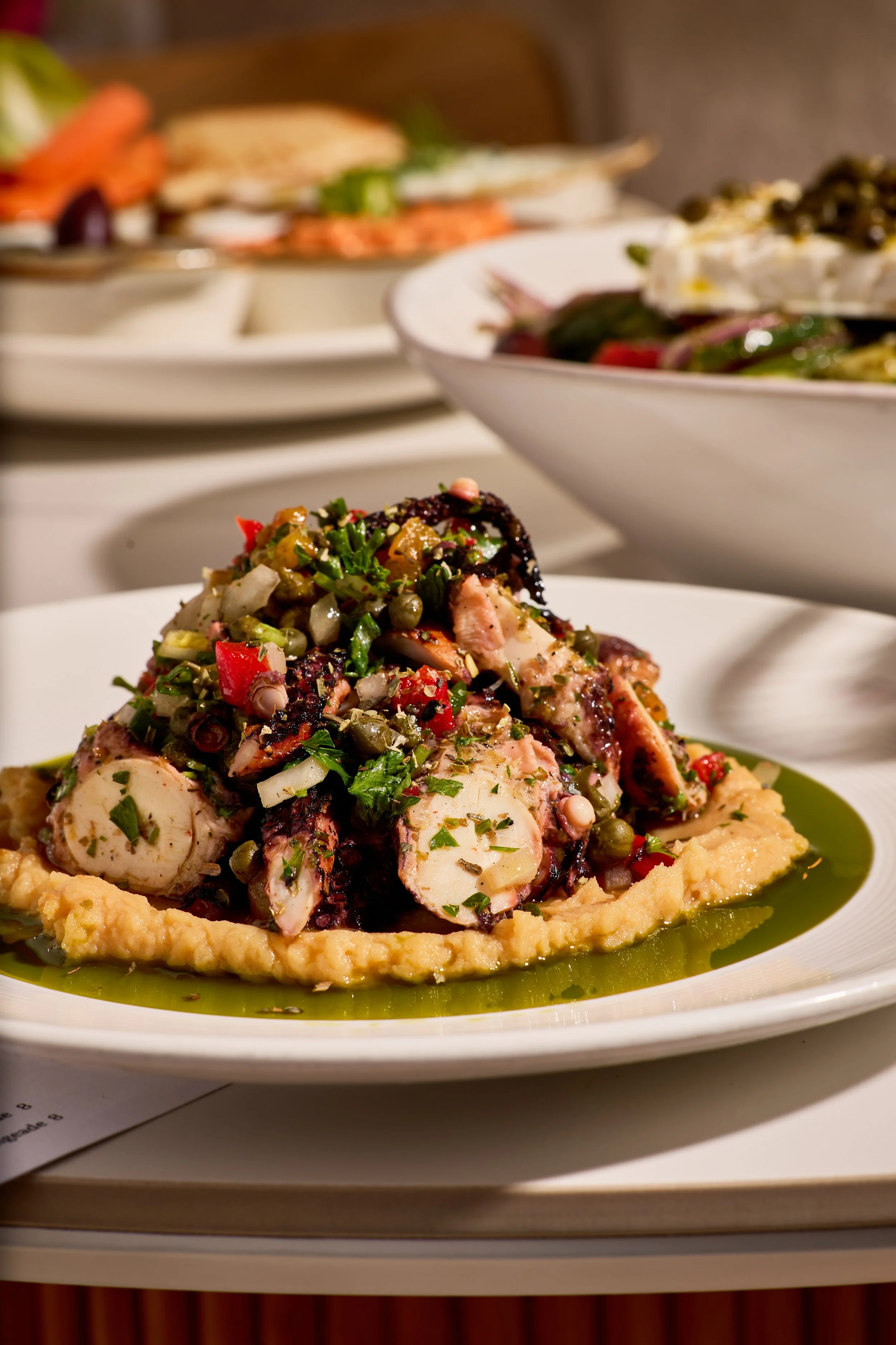 A plate of cooked octopus topped with chopped herbs, vegetables, and spices, served over a bed of mashed potatoes and olive oil.