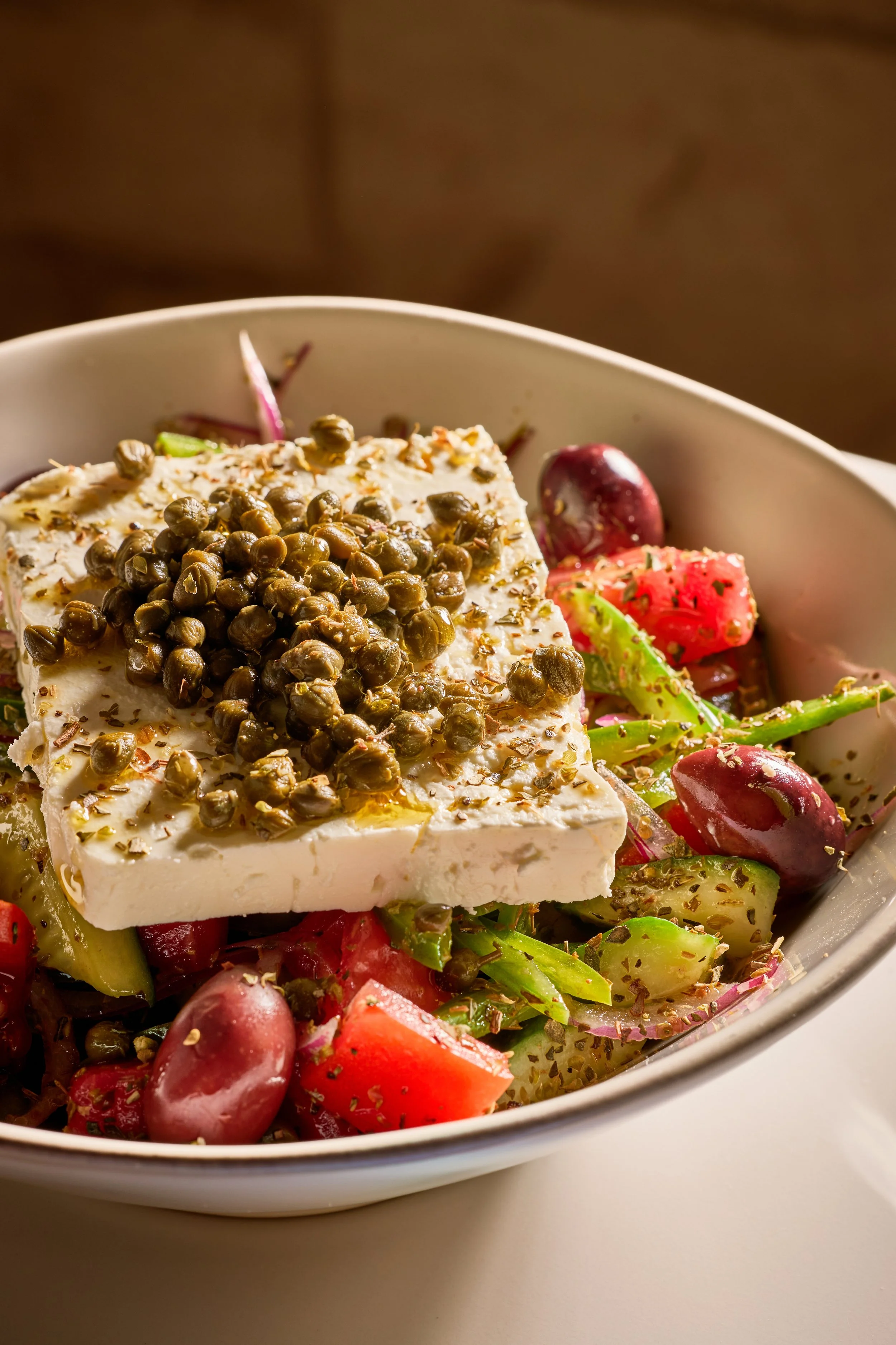 A bowl with a salad containing grape tomatoes, cucumber, and olives, topped with a large slice of feta cheese with capers and herbs.