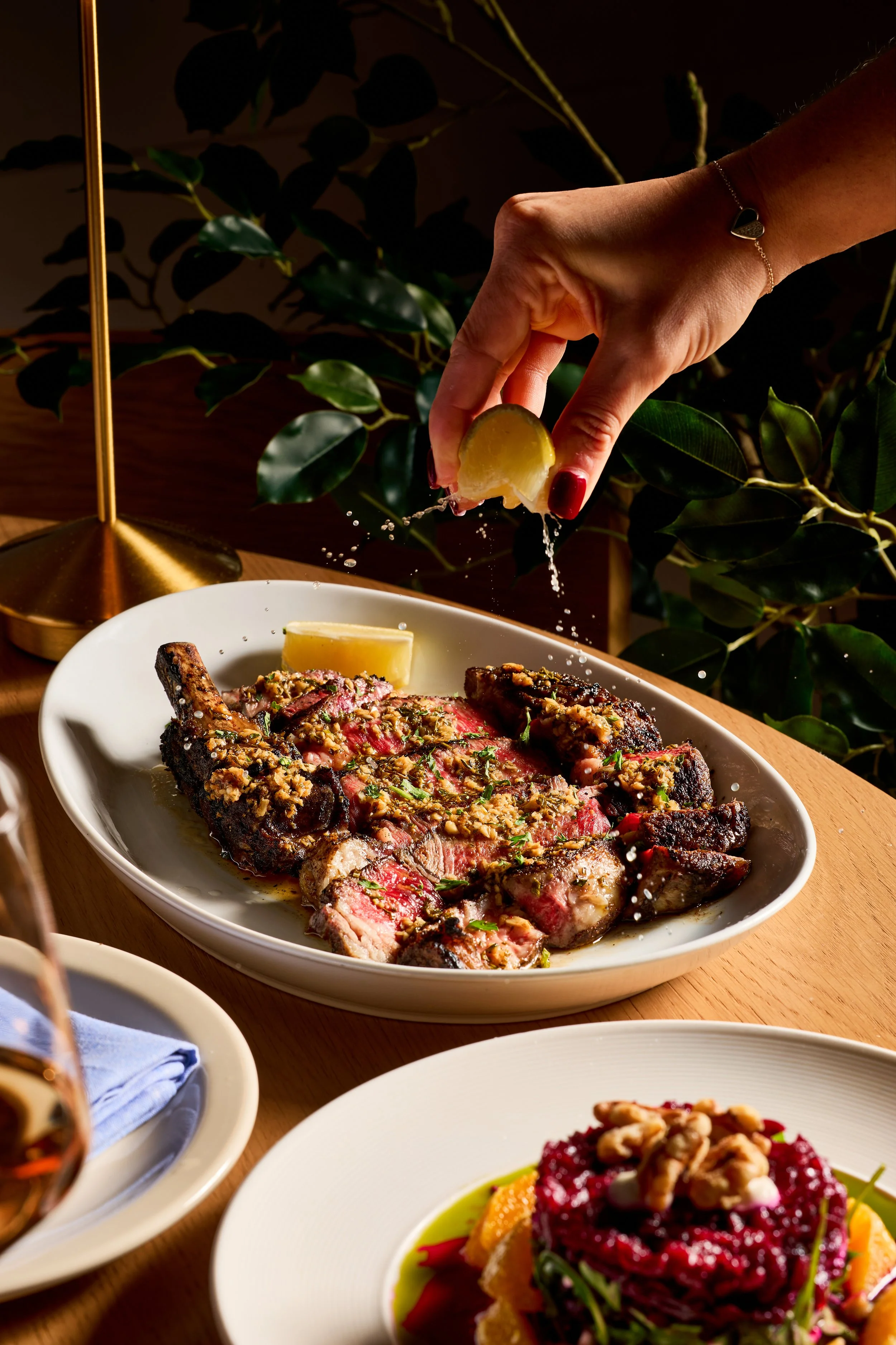 A hand squeezing a lemon over a plate of grilled steak with herbs, served with lemon wedges, on a wooden table with greenery in the background.