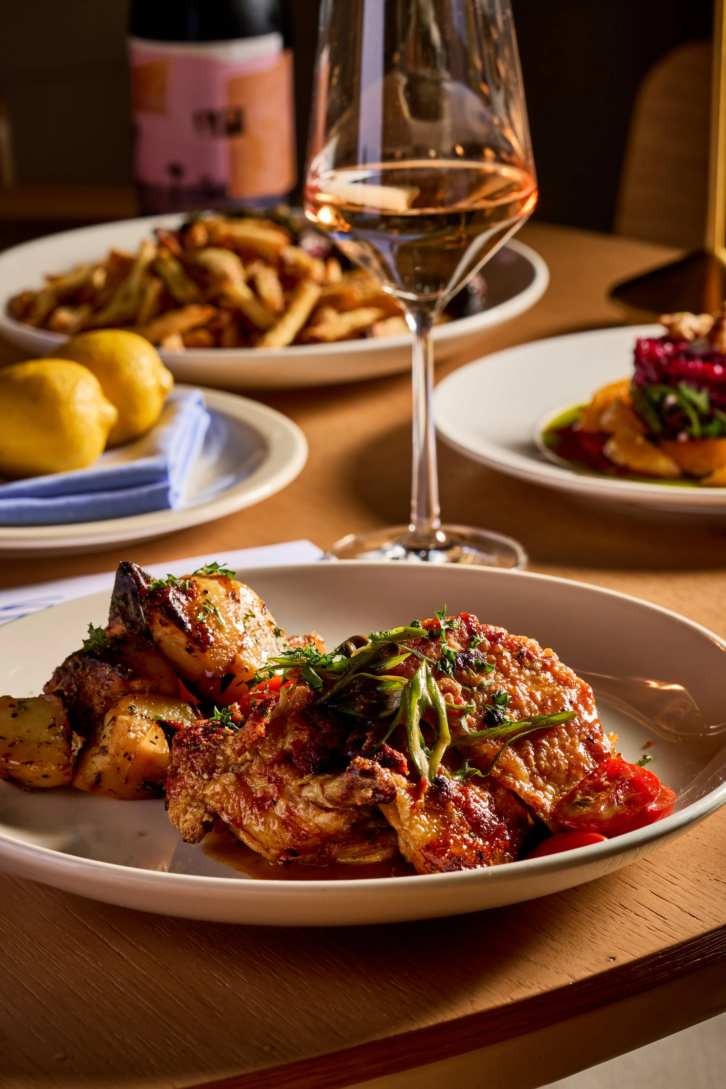 A plate of baked chicken with herbs, roasted potatoes, and cherry tomatoes on a wooden table, with a fork on the side, a glass of rosé wine, and other plates with food in the background.