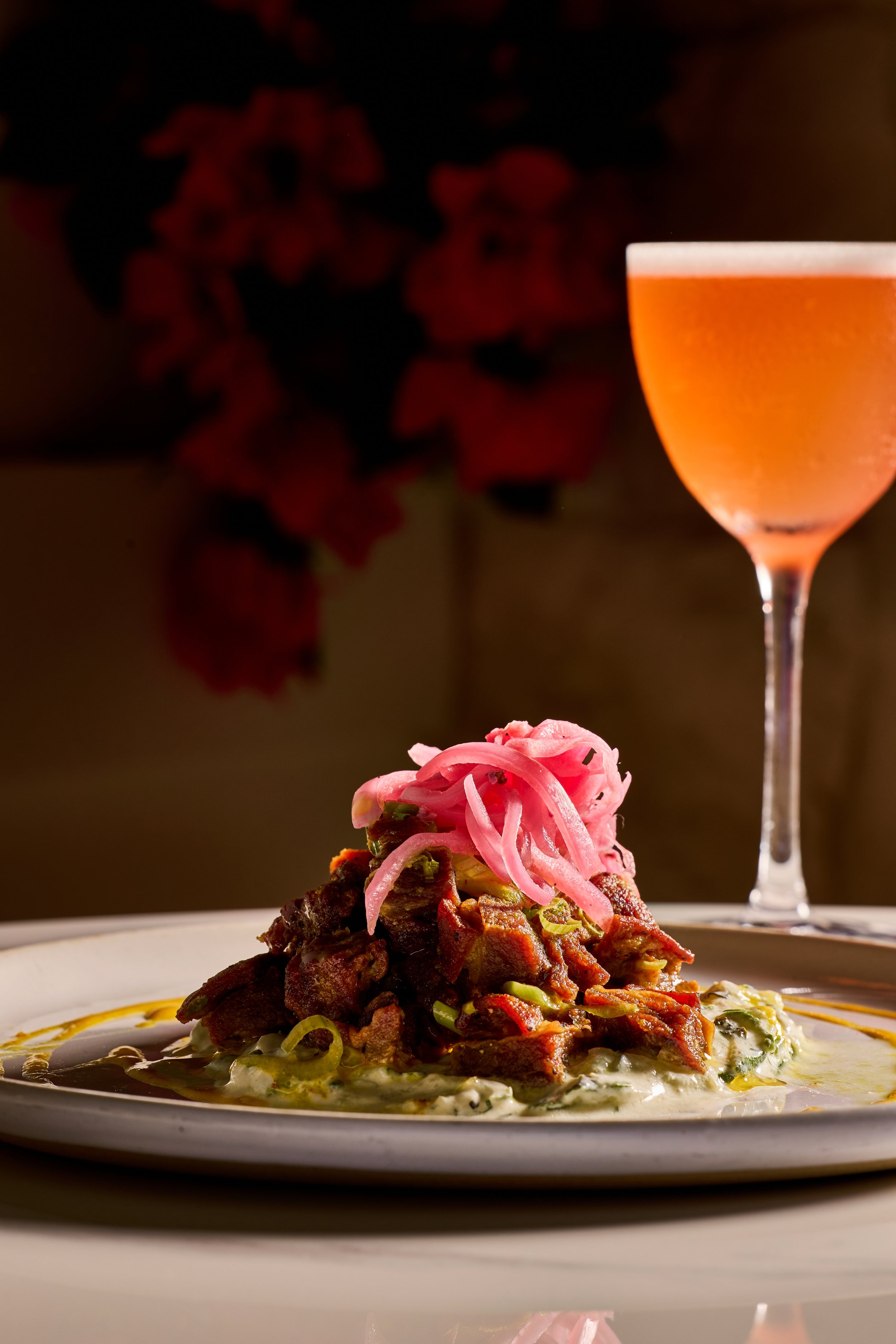 Plate of Mexican food with meat, onion, and green pepper, served with sauce, along with a pink cocktail in a tall glass, on a white table.