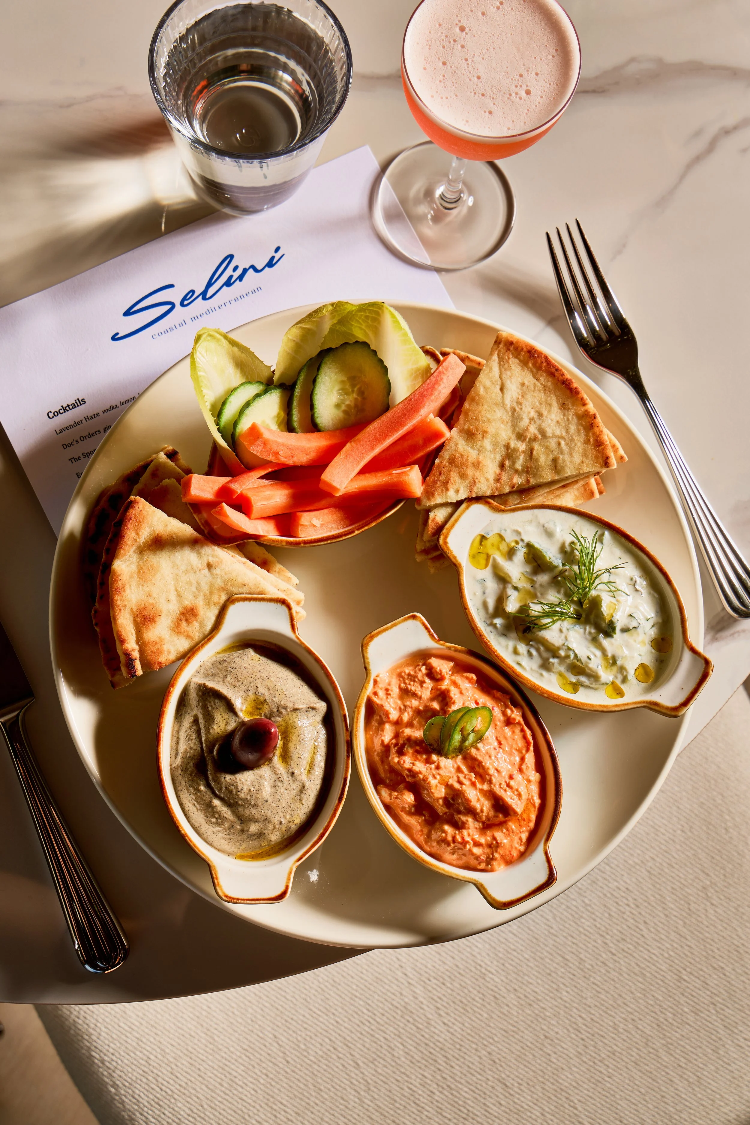 A plate of Mediterranean appetizers including tahini, hummus, and eggplant dip, served with pita bread, sliced cucumbers, lettuce, and pickled carrots, with a glass of water and a pink-frothed cocktail on the side.