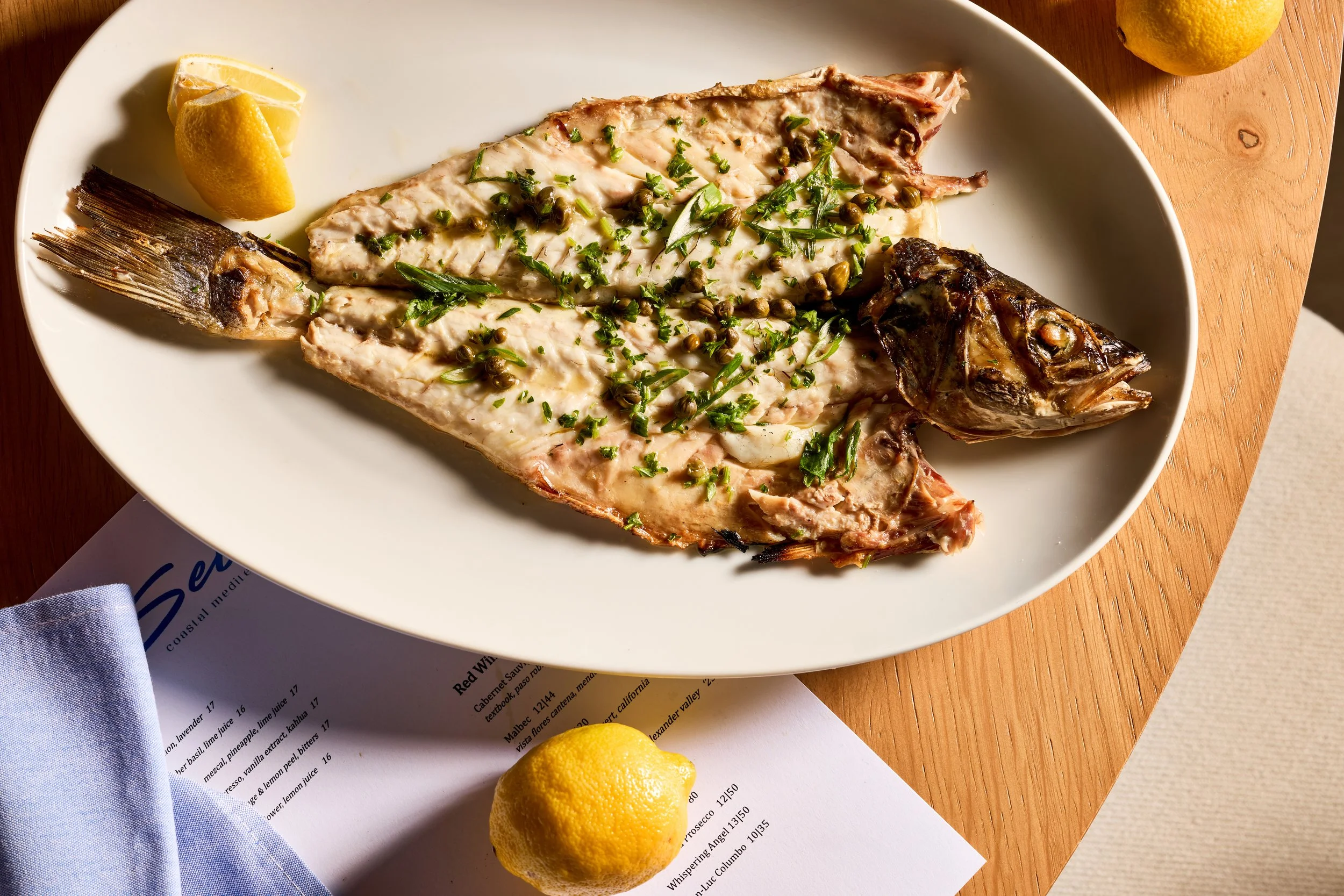 Whole grilled fish on a white oval plate garnished with chopped herbs and capers, accompanied by lemon wedges, placed on a wooden table with a lemon and a glass of water nearby.