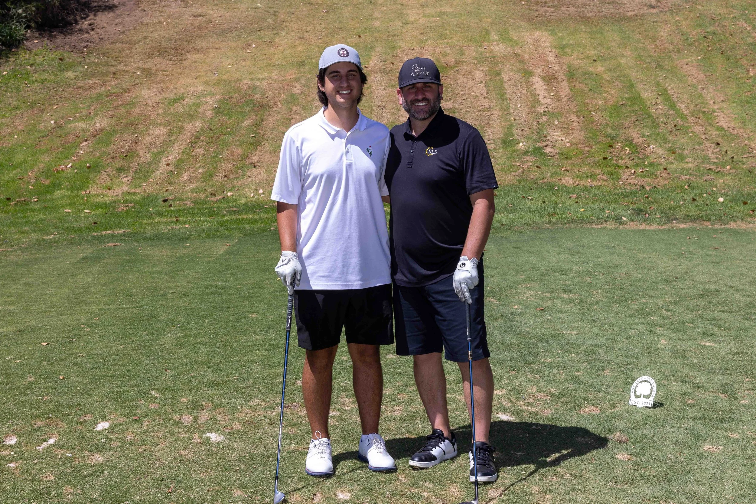Two men standing on a golf course, smiling, holding golf clubs, wearing golf attire and caps.