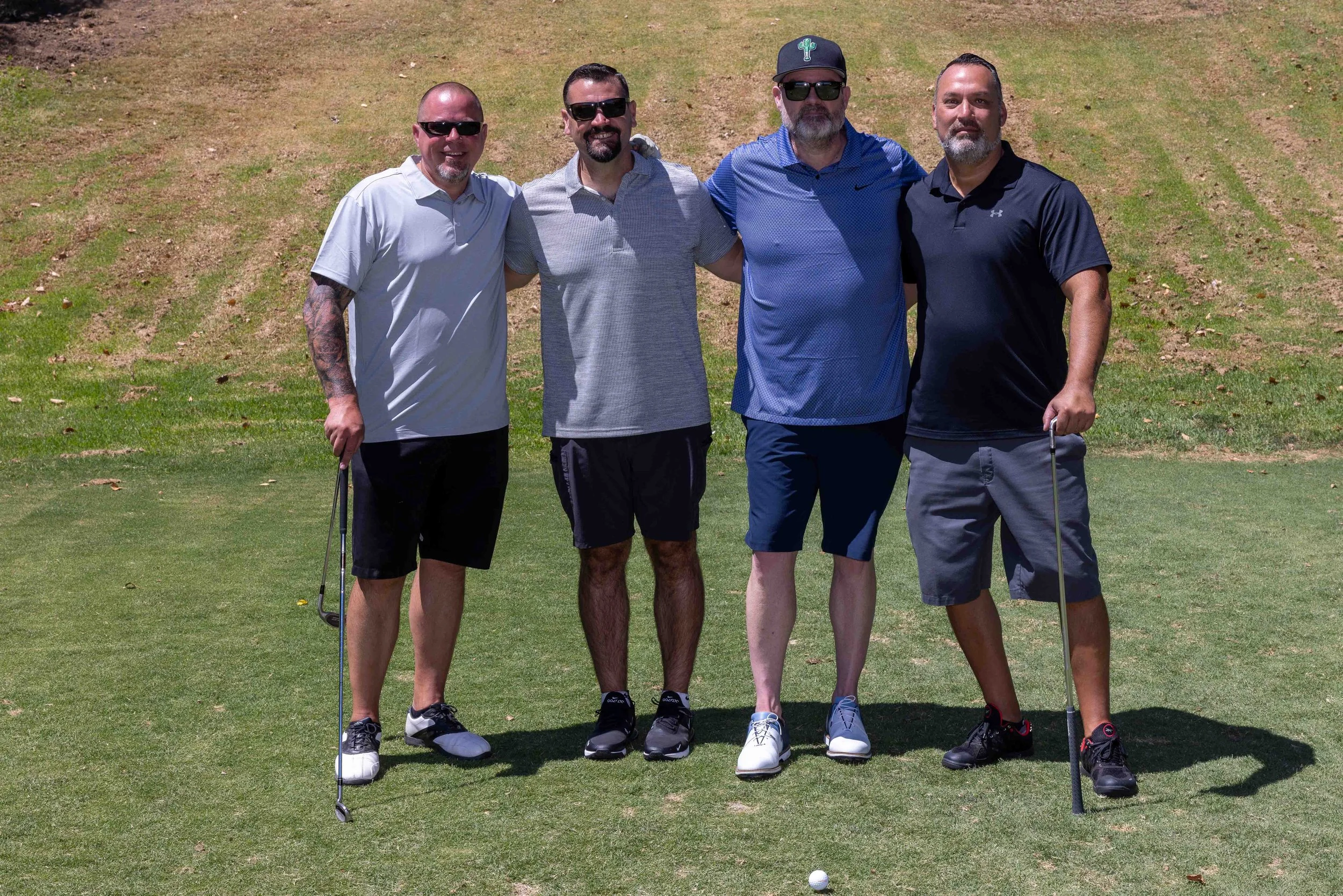 Four men standing on a golf course, posing for a photo, with one at the front holding a golf club and a golf ball on the ground.