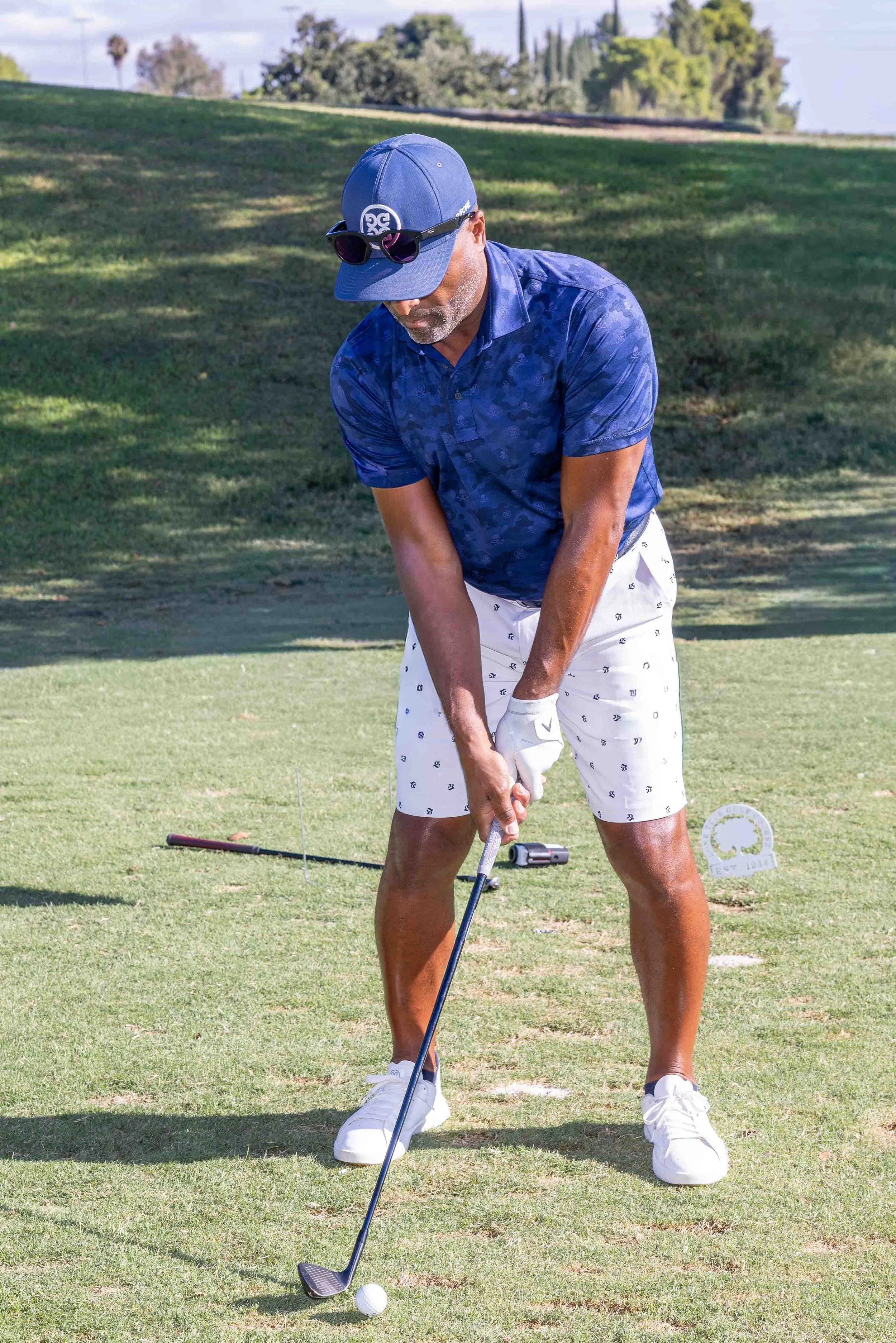 A man wearing a blue cap, sunglasses, a blue patterned polo shirt, and white shorts is preparing to swing a golf club on a golf course. There are trees and a scenic landscape in the background.