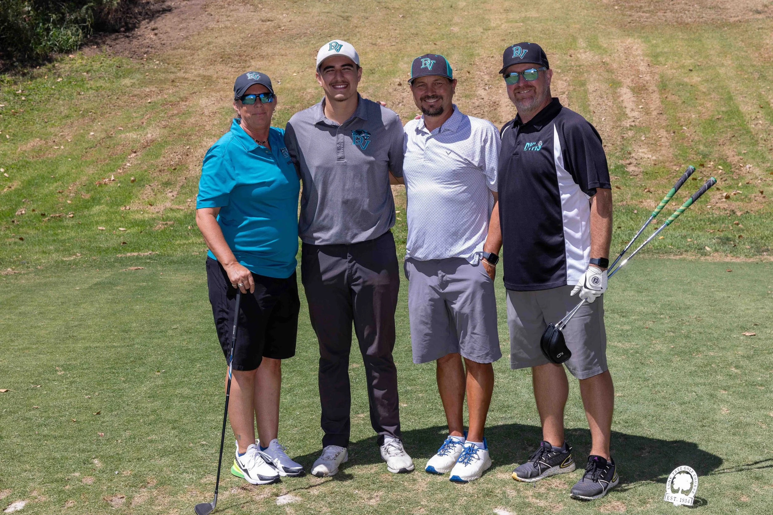 Four people standing on a golf course, smiling at the camera, with two holding golf clubs, dressed in golf attire, on a sunny day.