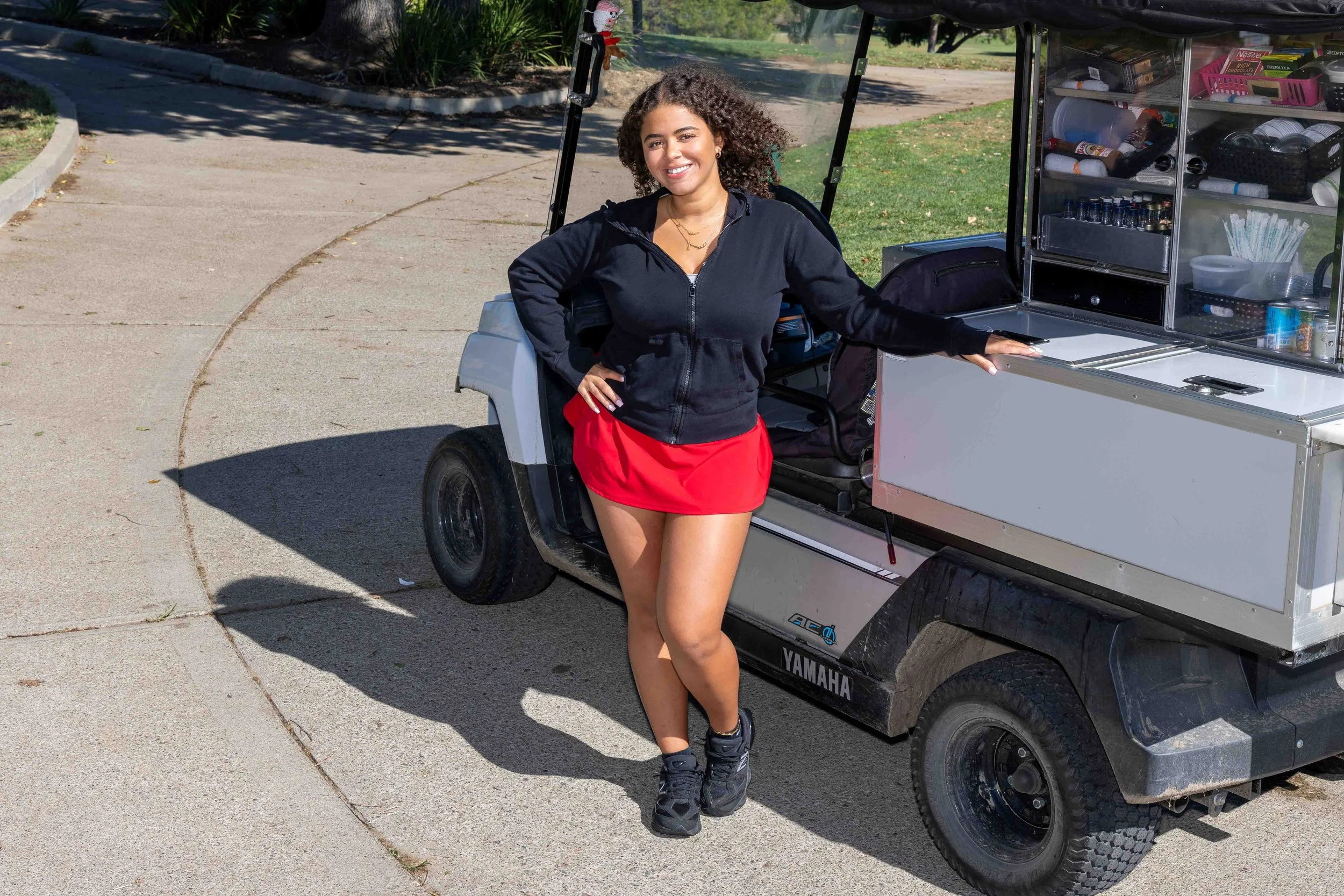 A young woman with curly hair smiling, wearing a black zip-up hoodie, red skirt, and athletic shoes, standing beside a small utility vehicle with a snack and drink cart on it in an outdoor park setting.