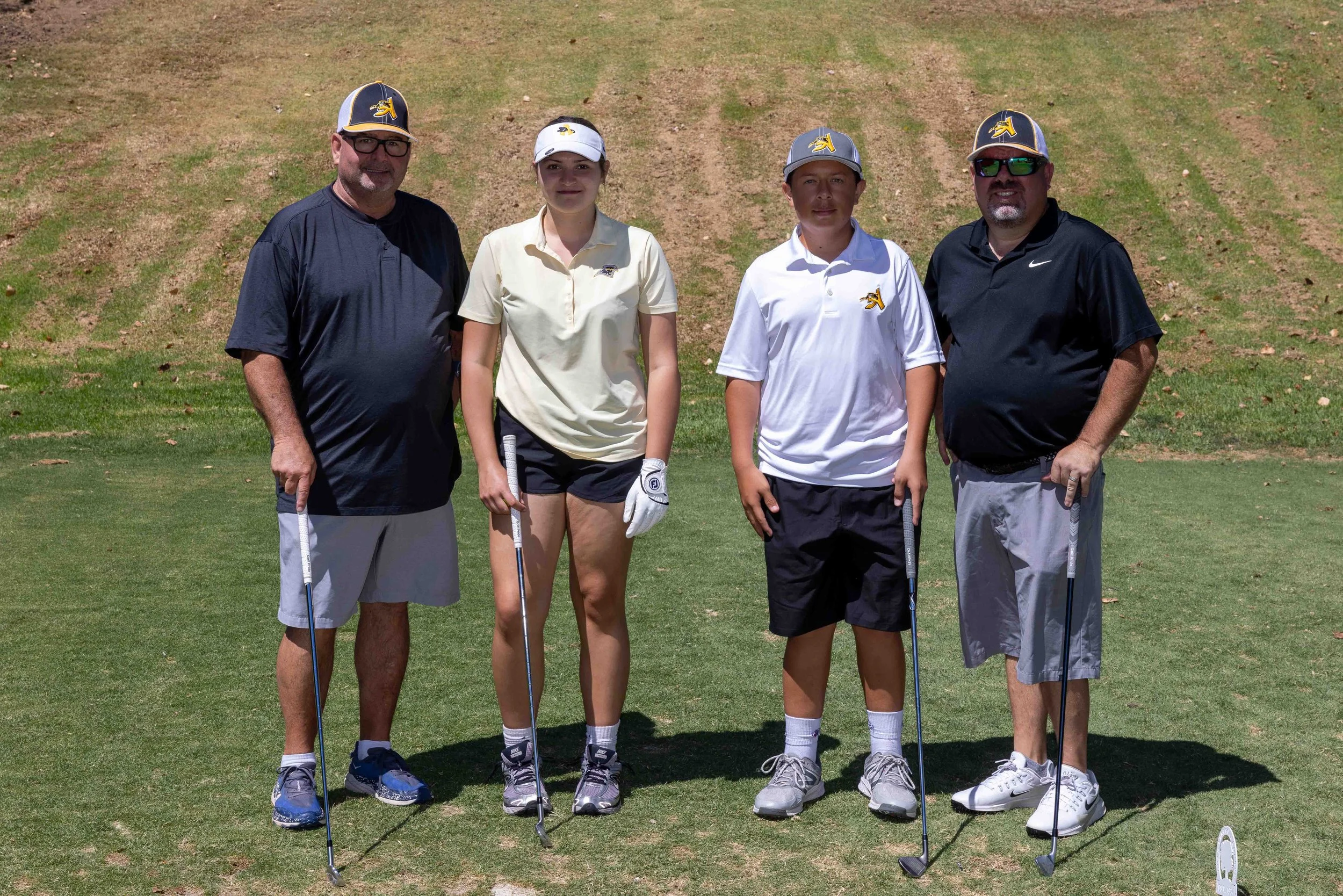 Group of four people on a golf course, holding golf clubs, wearing golf caps and athletic clothing, posing for a photo.