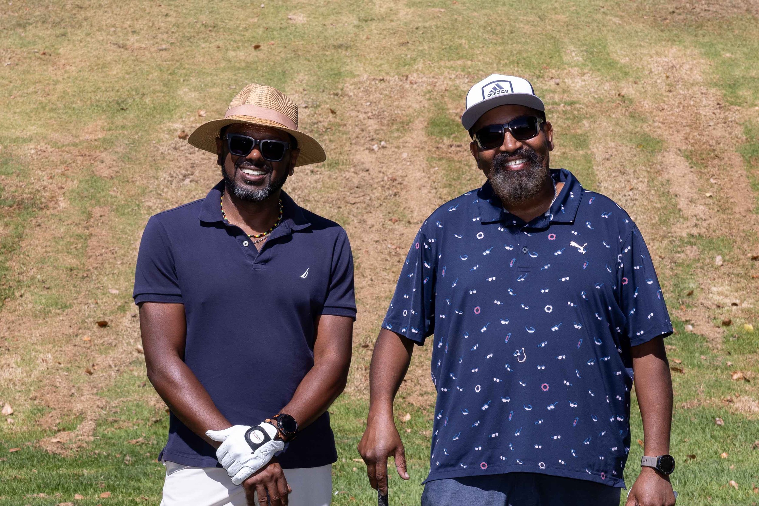 Two men with dark skin smiling outdoors on a sunny day, wearing sunglasses and hats, standing on a grassy area with patches of dirt.