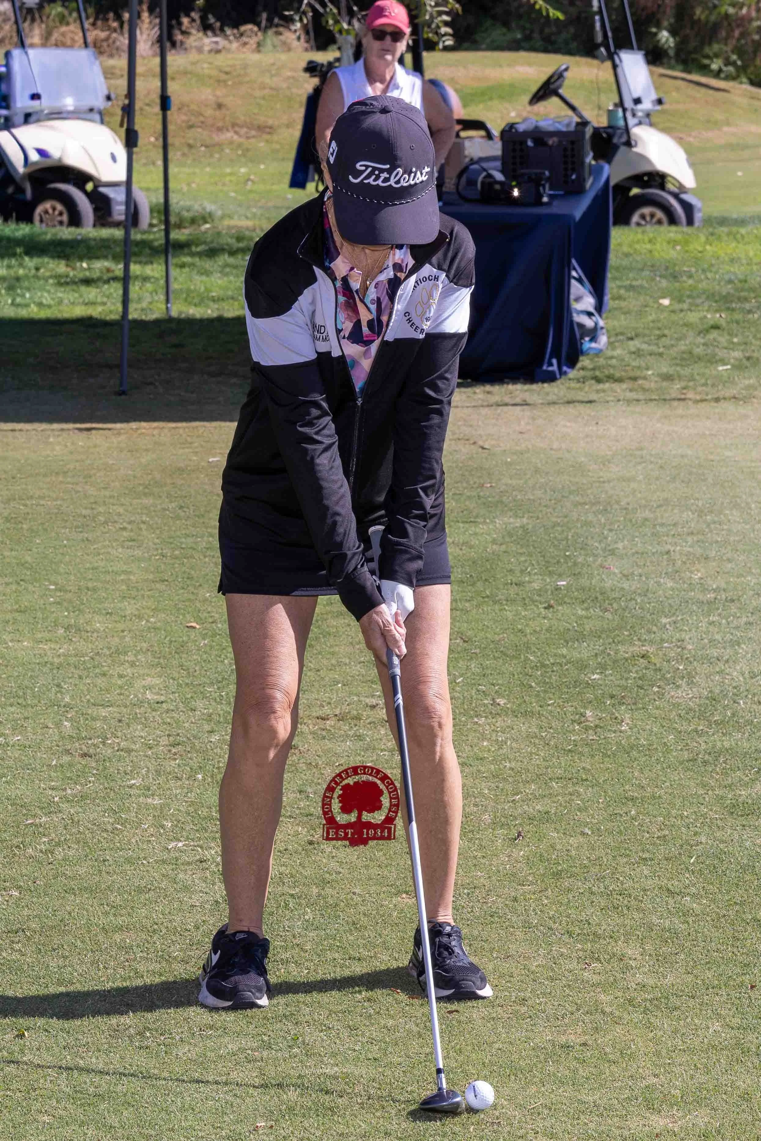 A woman prepares to tee off on a golf course with a golf club, wearing a black cap, black jacket, colorful shirt, and black shoes. Another woman stands in the background near golf carts and equipment.