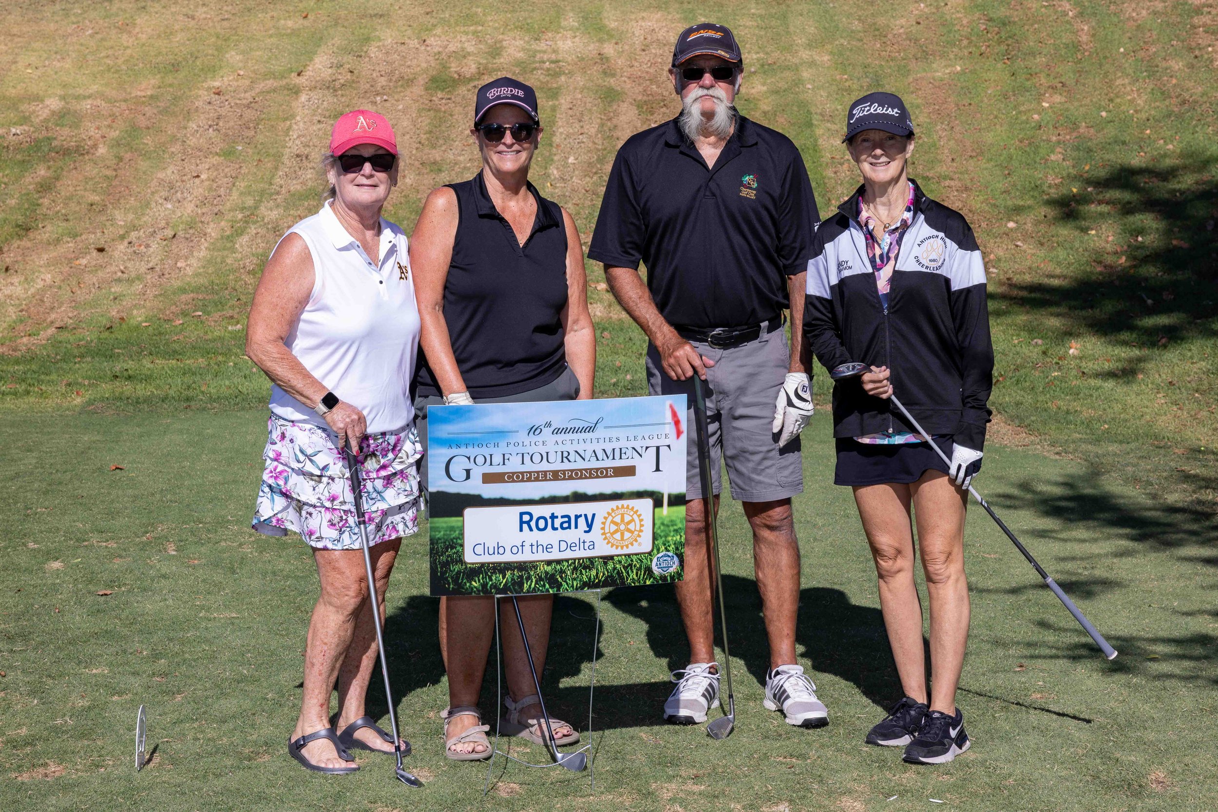Four women and one man standing on a golf course, holding golf clubs, with a sign that reads '16th annual Antioch Police Activities League Golf Tournament, Copper Sponsor, Rotary Club of the Delta.' They are dressed in golf attire and are smiling for