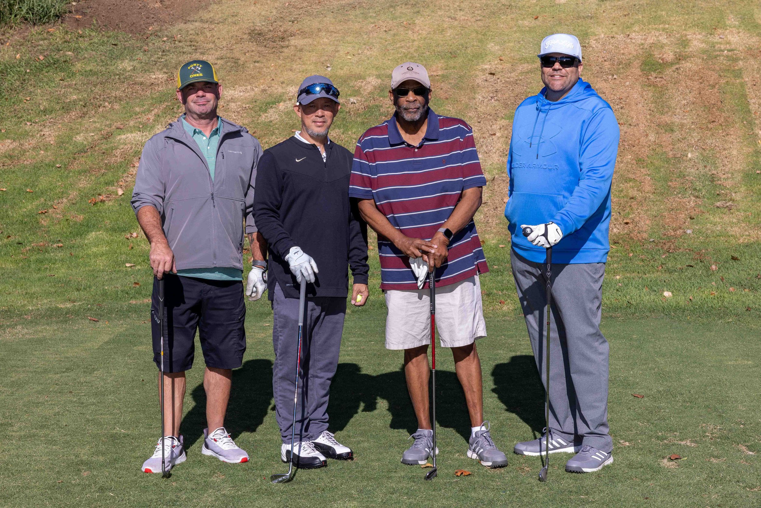 Four men standing on a golf course holding golf clubs, dressed in casual golf attire, with a grassy slope in the background.