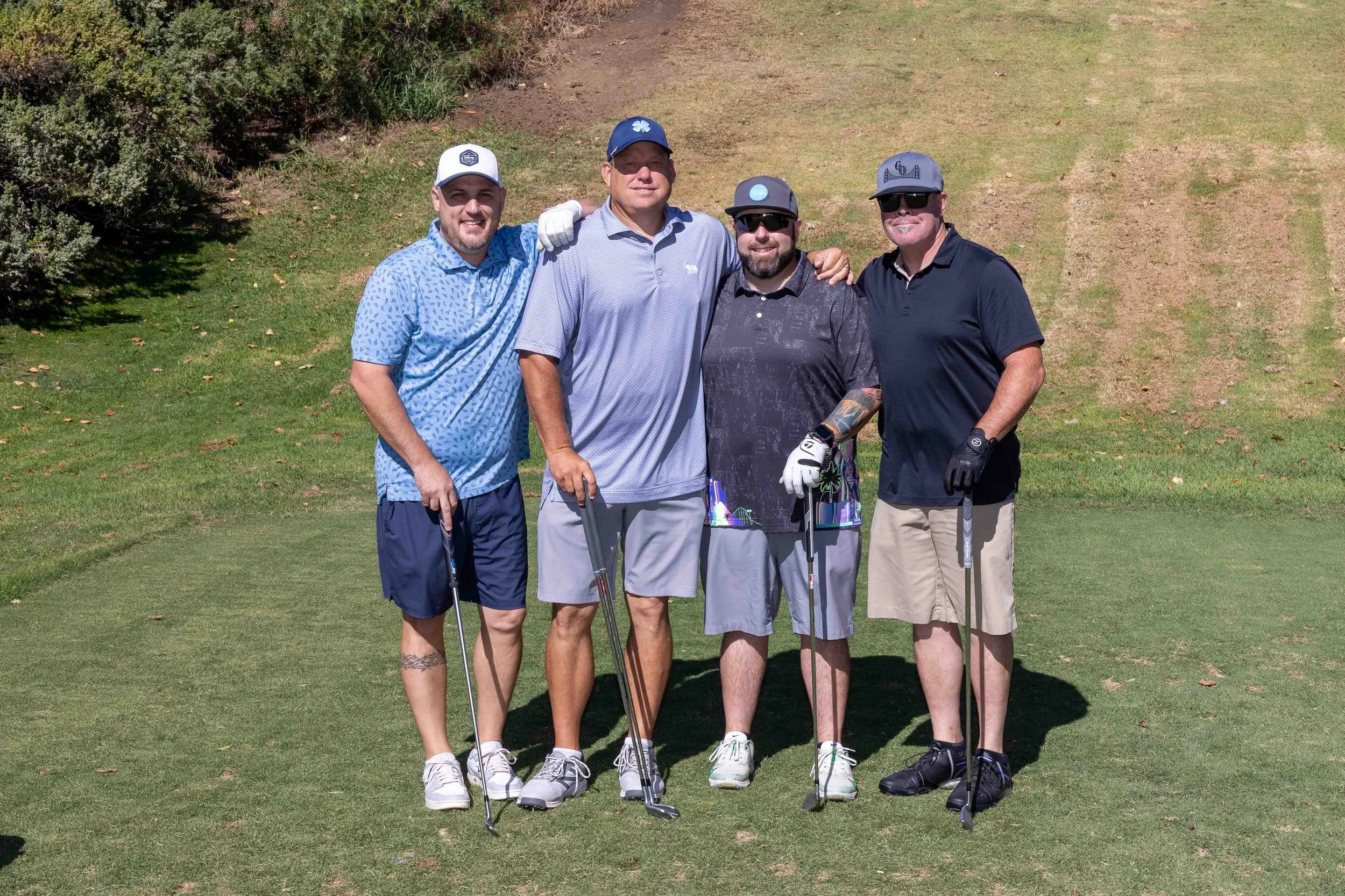 Four men standing on a golf course, smiling at the camera. They are dressed in golf attire, holding golf clubs, and appear to be enjoying a sunny day.