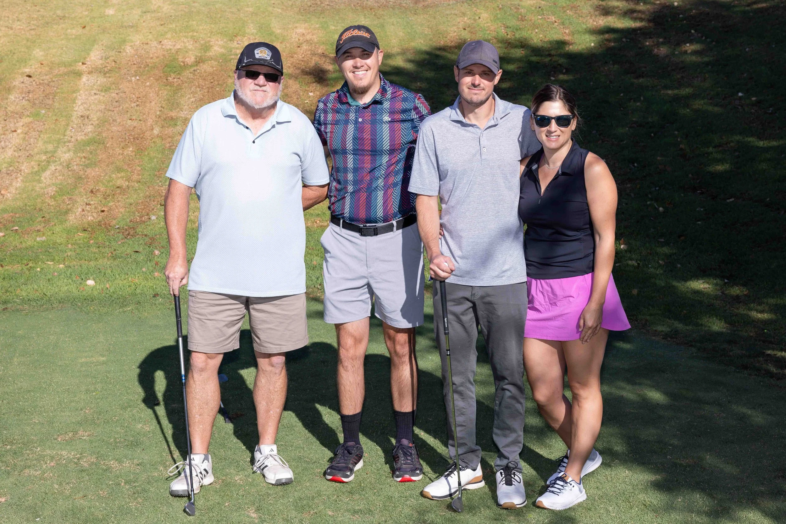Group of five people standing on a golf course with two holding golf clubs, smiling at the camera, wearing casual golf attire and sunglasses, with trees and grass in the background.