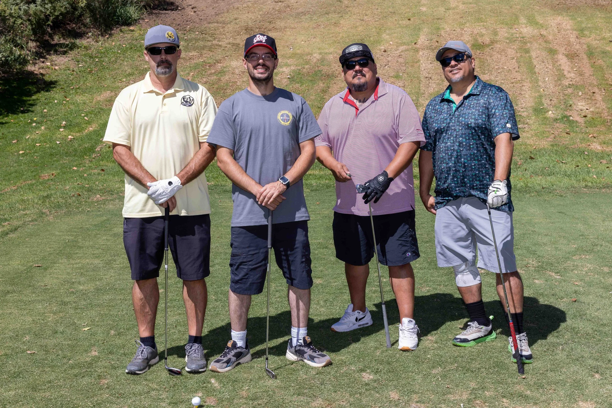 Four men standing on a golf course holding golf clubs, wearing sunglasses, casual golf attire, and smiling.