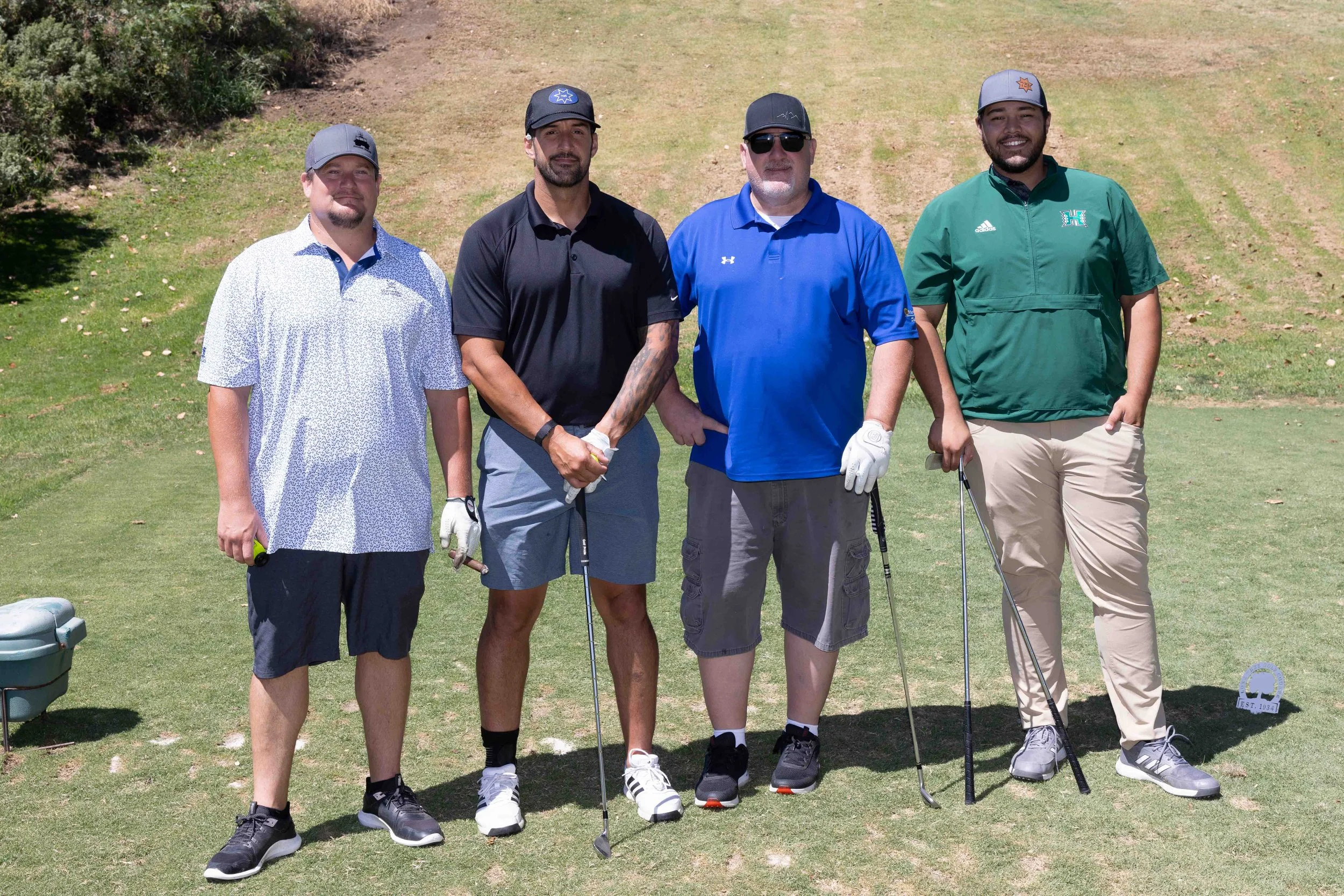 Four men posing on a golf course, holding golf clubs and wearing casual golf attire, standing in front of a grassy hill with trees to the side.