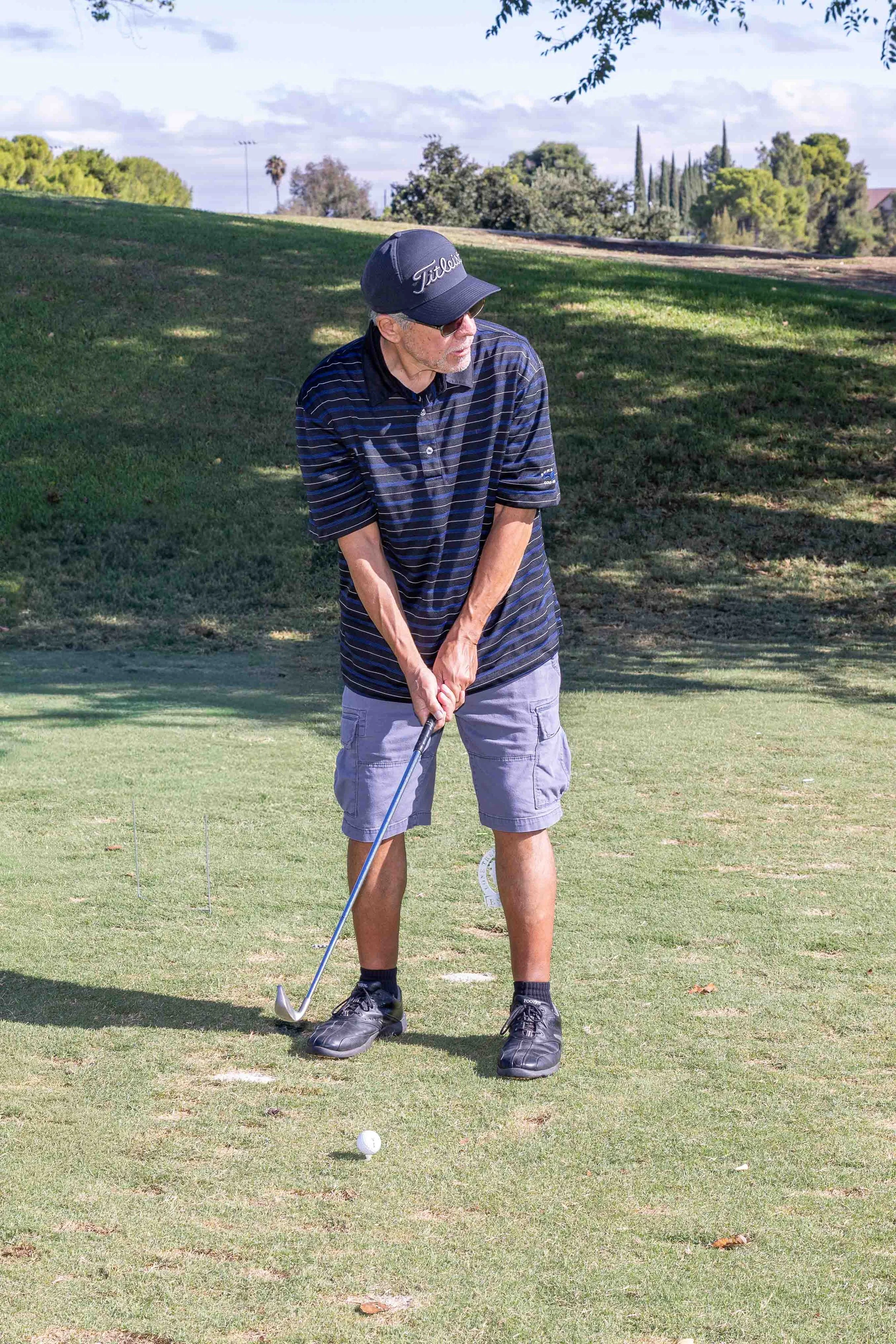 A man playing golf on a sunny day, preparing to hit a golf ball with a club on a well-maintained golf course surrounded by trees.