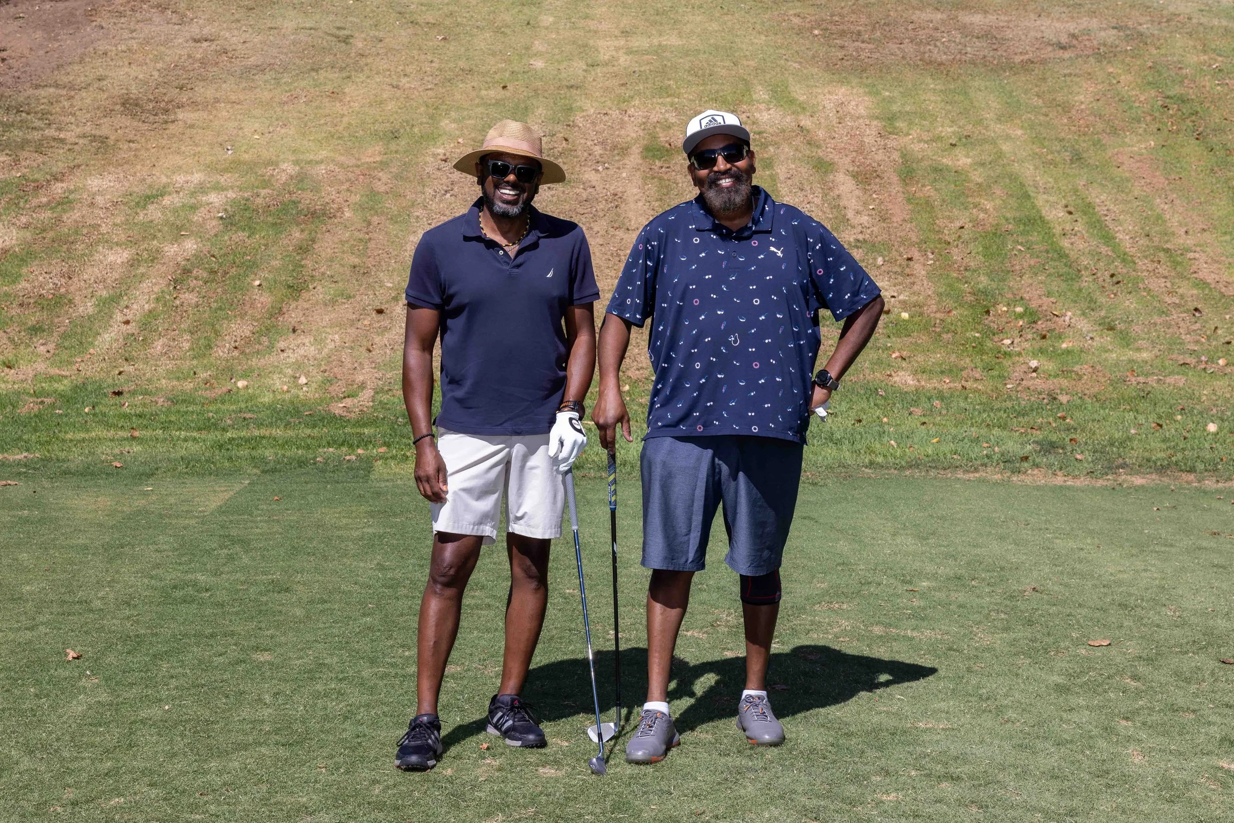 Two men standing on a golf course, smiling, with one holding a golf club. They are dressed in casual summer golf attire, wearing sunglasses and hats, with a grassy and slightly dry landscape behind them.