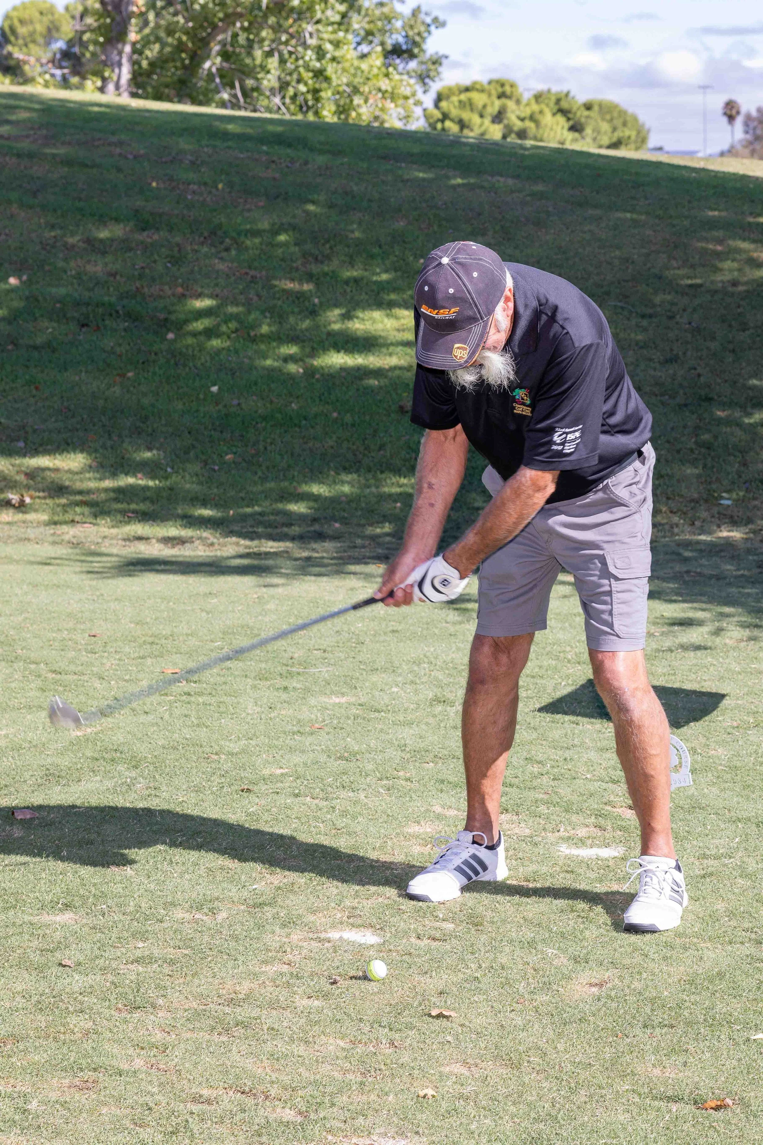 A man with a beard and glasses playing golf on a sunny day, wearing a black cap, black polo shirt, light gray shorts, and white shoes, about to hit a golf ball on the grass.