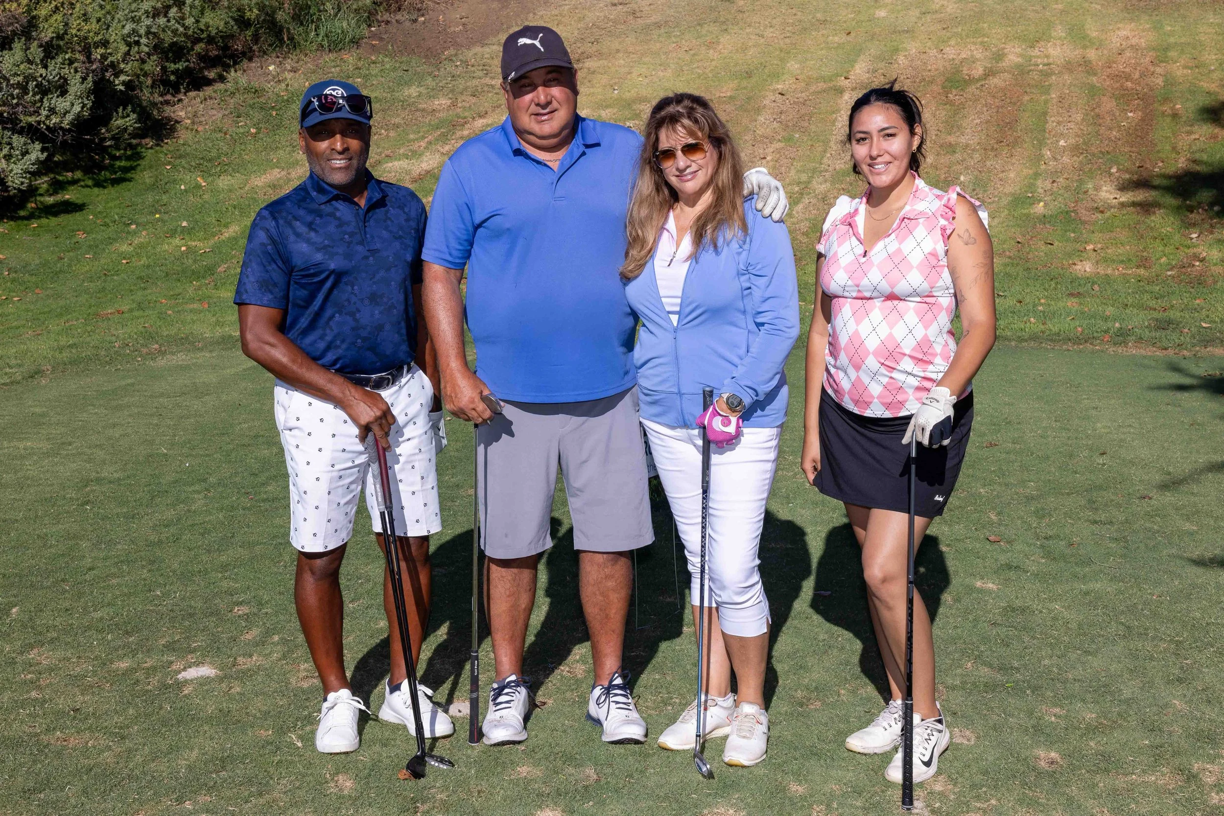 Four people standing on a golf course, holding golf clubs, dressed in sporty casual clothing, smiling at the camera.