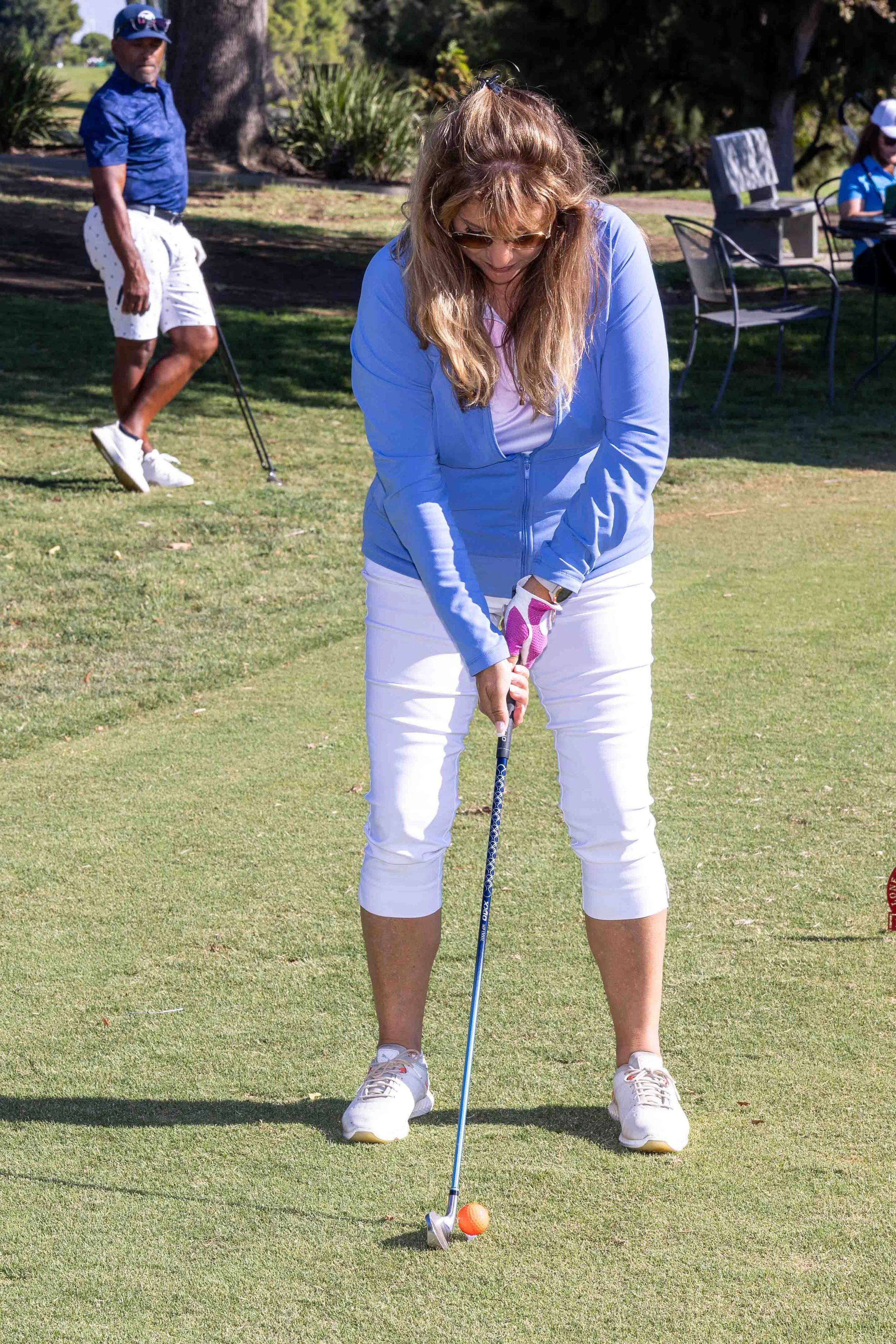 A woman wearing a light blue jacket, white pants, and white sneakers is preparing to hit a golf ball on a golf course. She is wearing sunglasses and has long, blonde hair. In the background, a man in a blue shirt, white shorts, and a cap is standing 