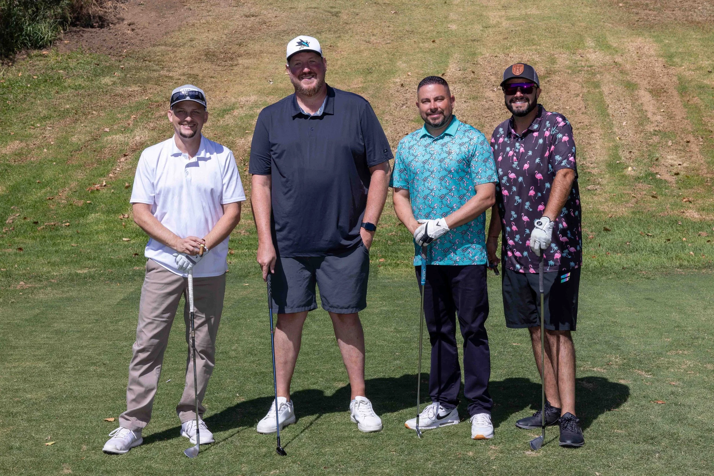Group of four men standing on a golf course, holding golf clubs, smiling at the camera, wearing casual golf attire including hats and sunglasses.